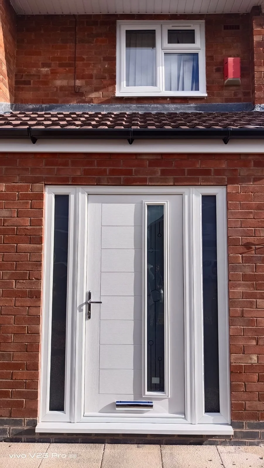 Front door of a red brick house with white door and side windows, upper window with curtains, and roof with tiles.
