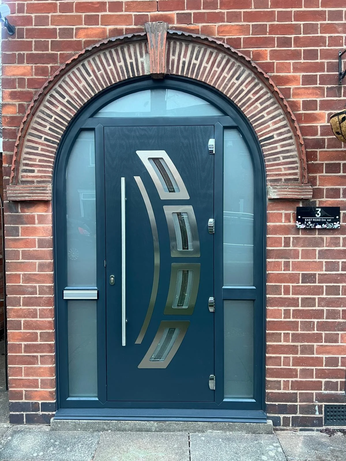 Modern blue front door with geometric silver design, flanked by frosted glass panels, set in a brick wall with an arched brick lintel above it.