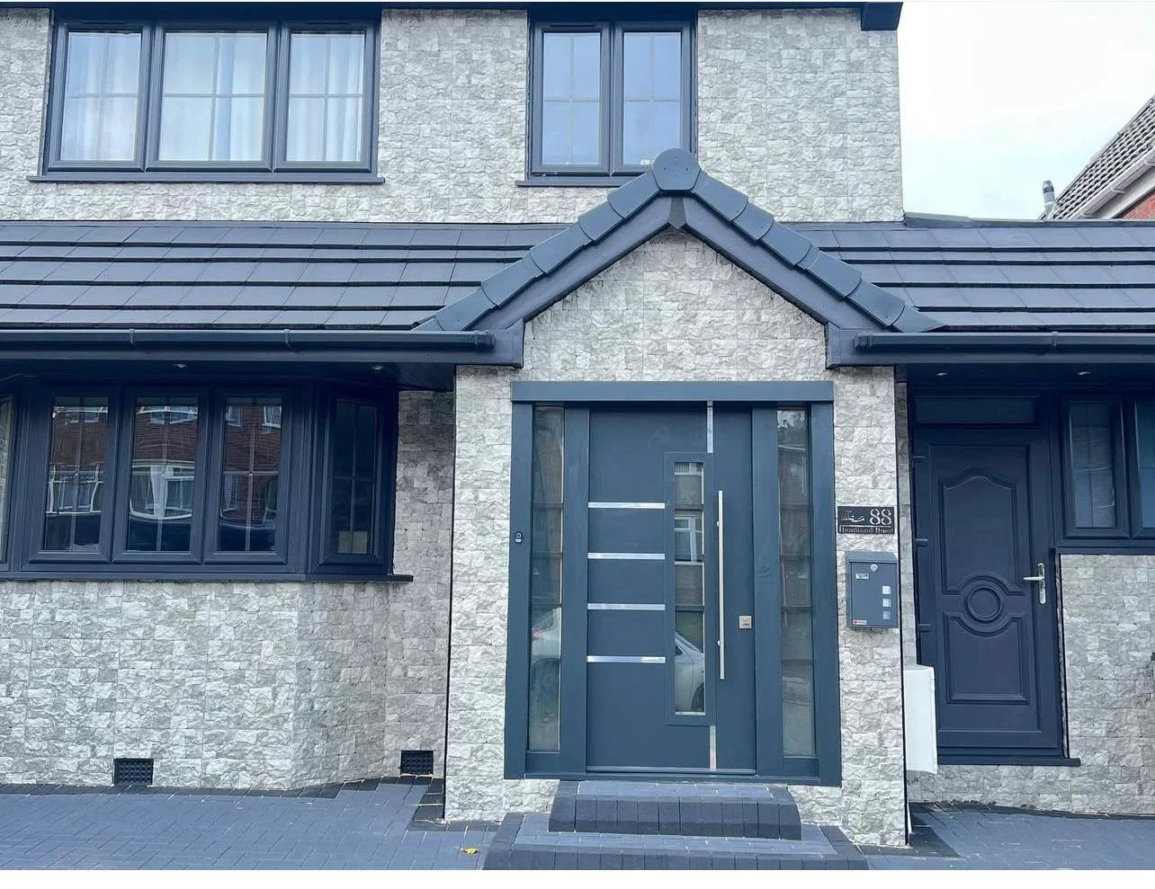 Front view of a modern house with a stone exterior, black-framed windows, a front door with vertical glass panels, and a house number sign.