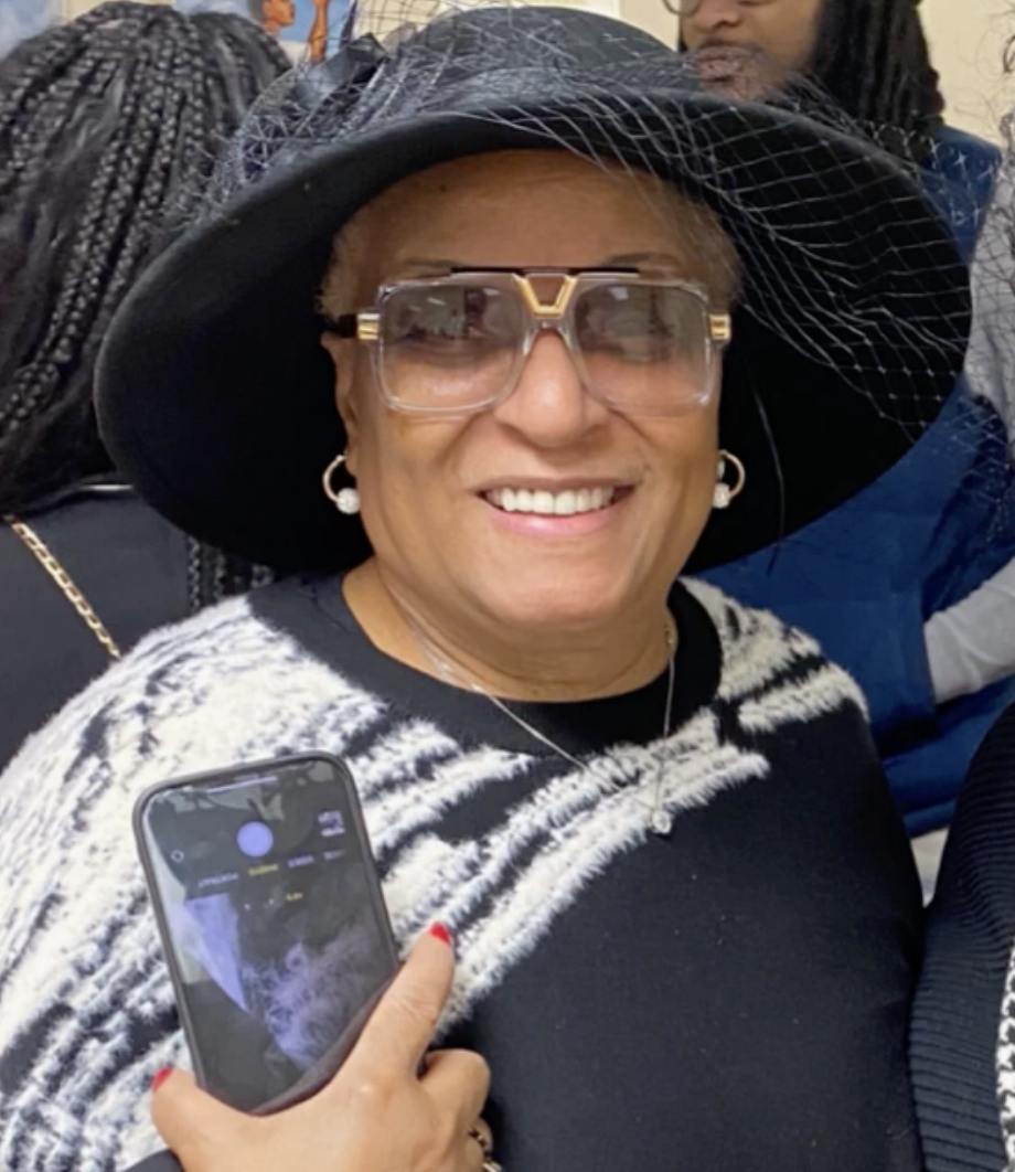 A smiling woman wearing a large black hat with netting, clear glasses with gold accents, earrings, and a black and white patterned sweater, holding a smartphone with a camera app open.