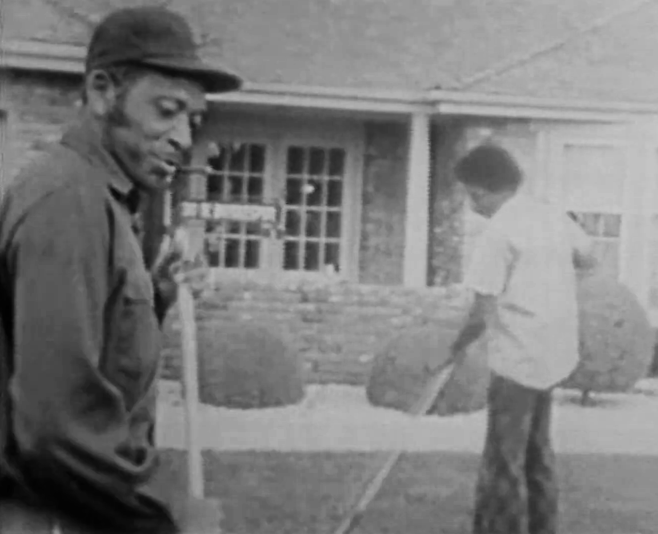 Black and white photo of two men standing outdoors near a building. One man on the left is smoking a cigar and wearing a cap. The other man on the right is carrying a bag.
