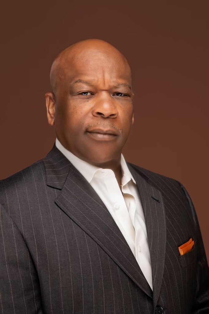 A bald African American man wearing a black pinstripe suit with a white shirt and an orange pocket square, posing against a brown background.