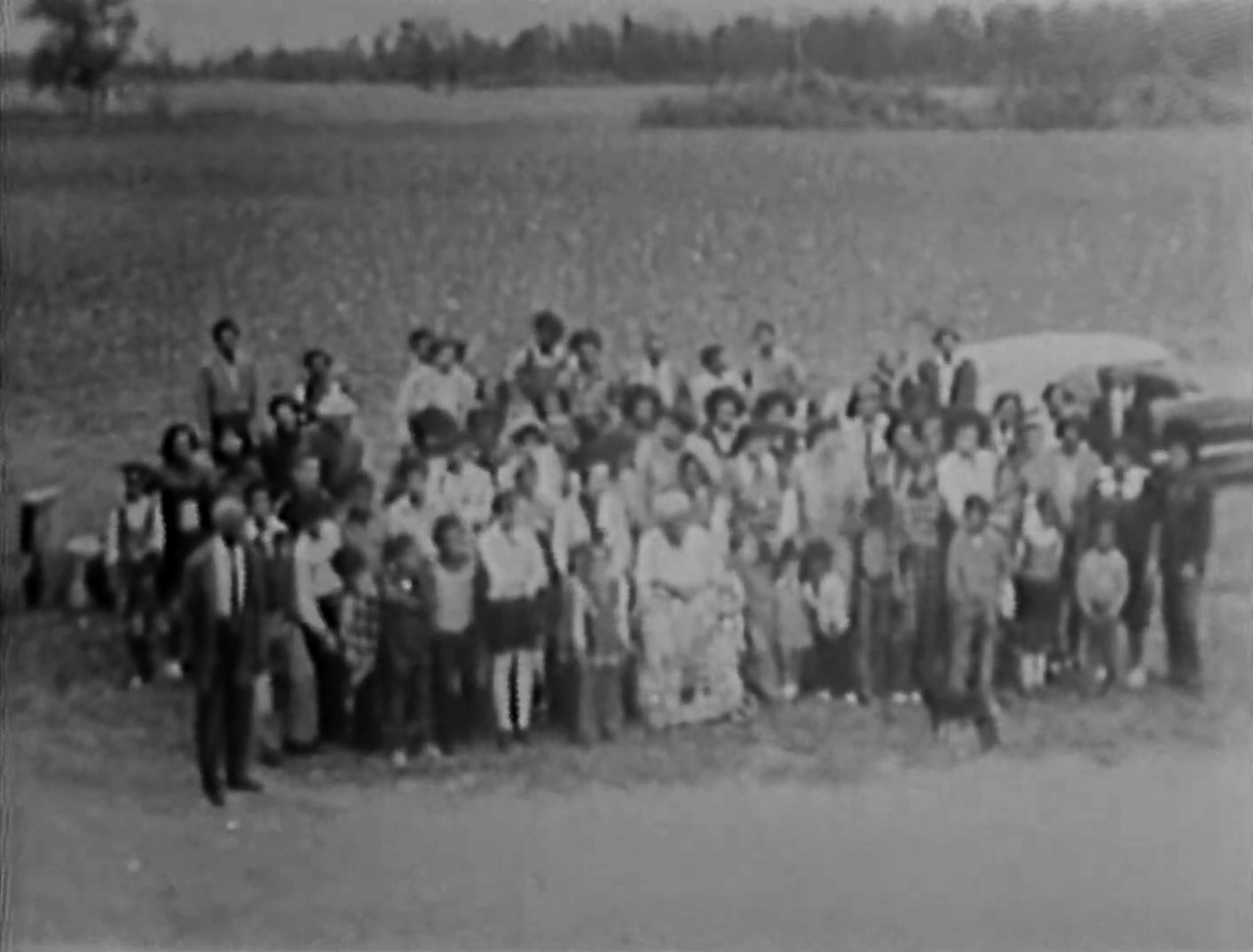 A large group of people, including children and adults, gathered outdoors in a field, with some standing and some sitting, possibly posing for a photo. There are trees and open land in the background, and a covered vehicle is visible on the right side.