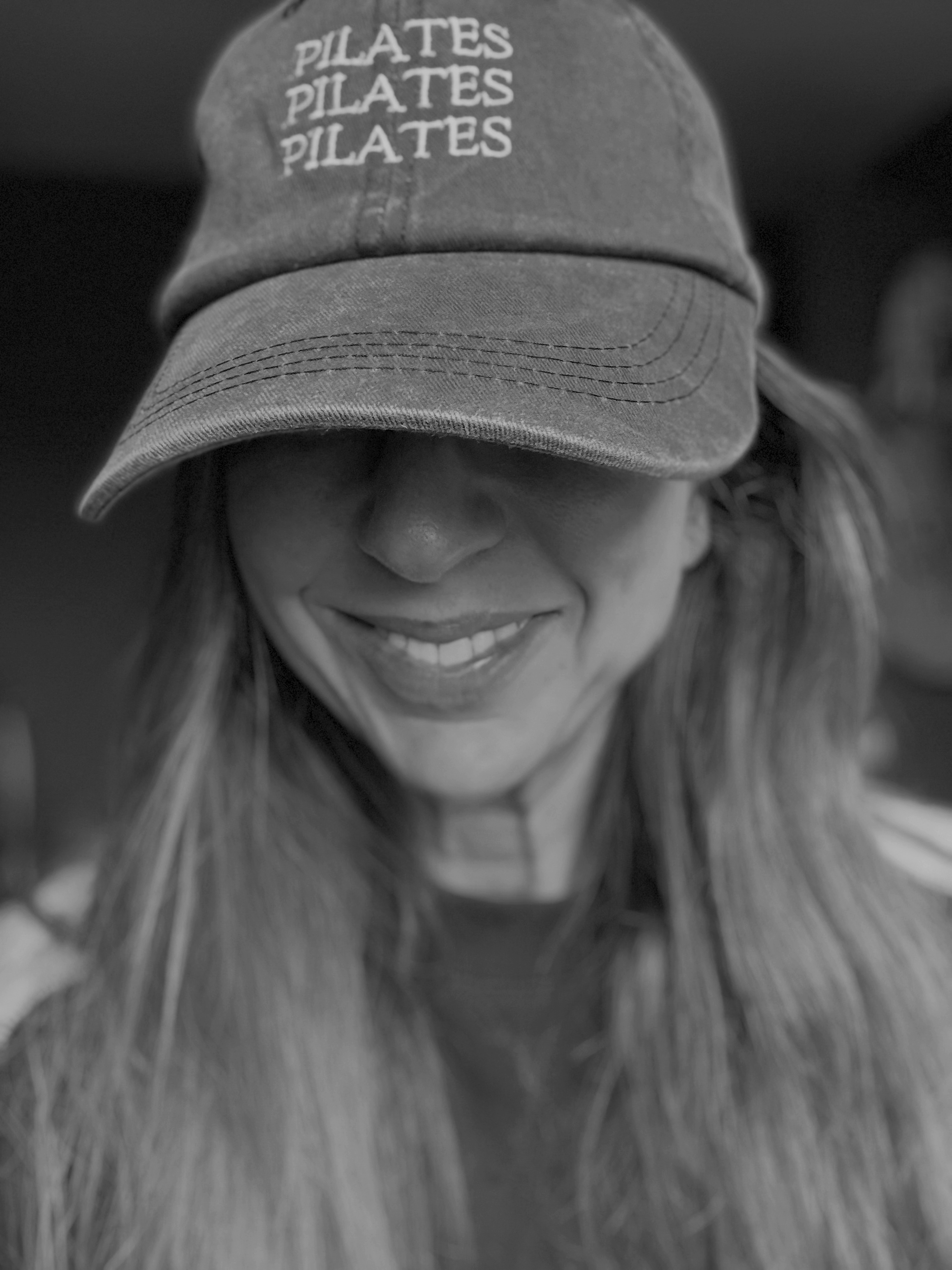 A woman with long hair smiling wearing a cap that says 'Pilates' multiple times.