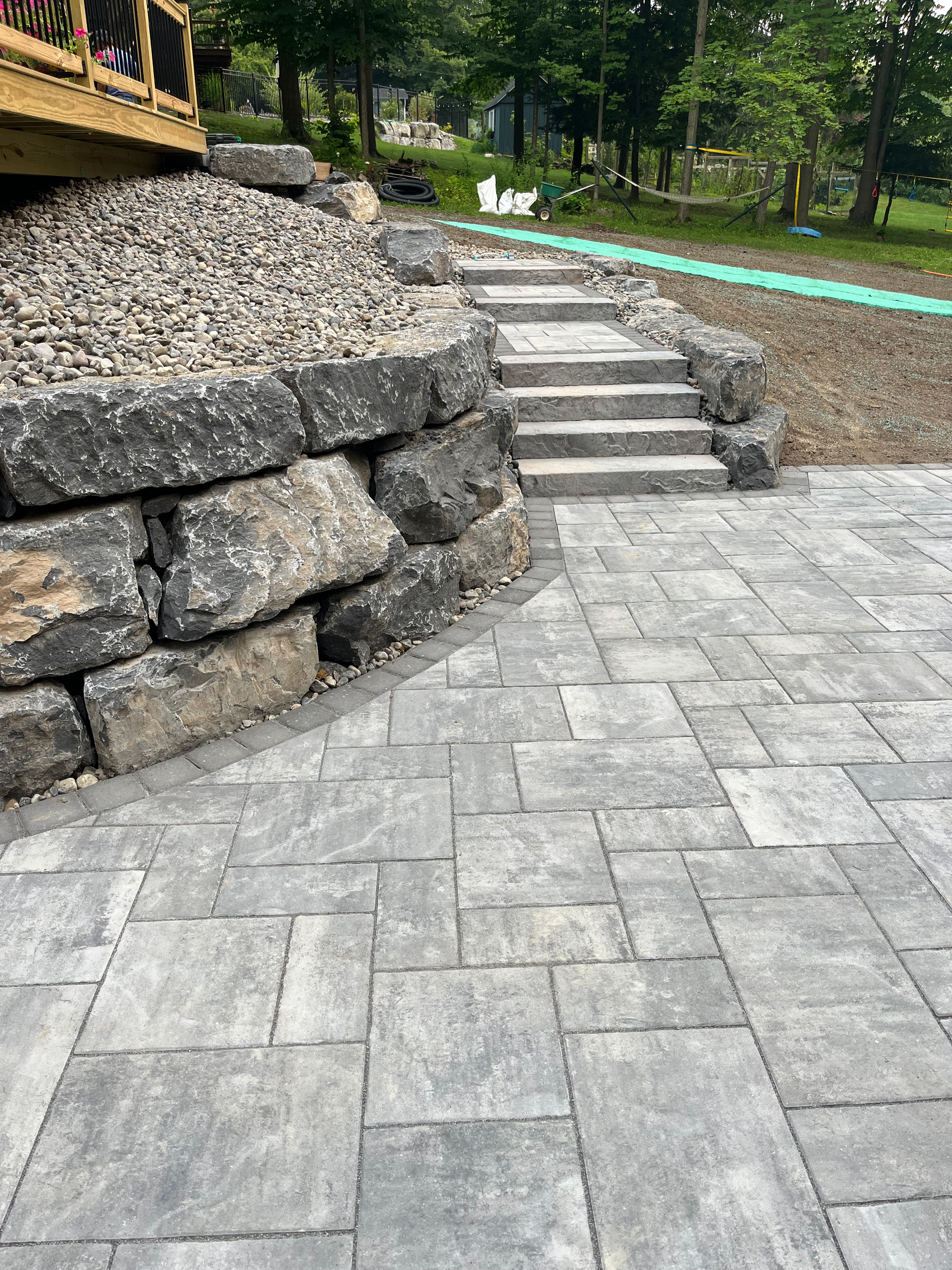 Stone retaining wall with large rocks and small pebbles, concrete steps leading up, and a paved stone patio in a backyard with trees and grass.