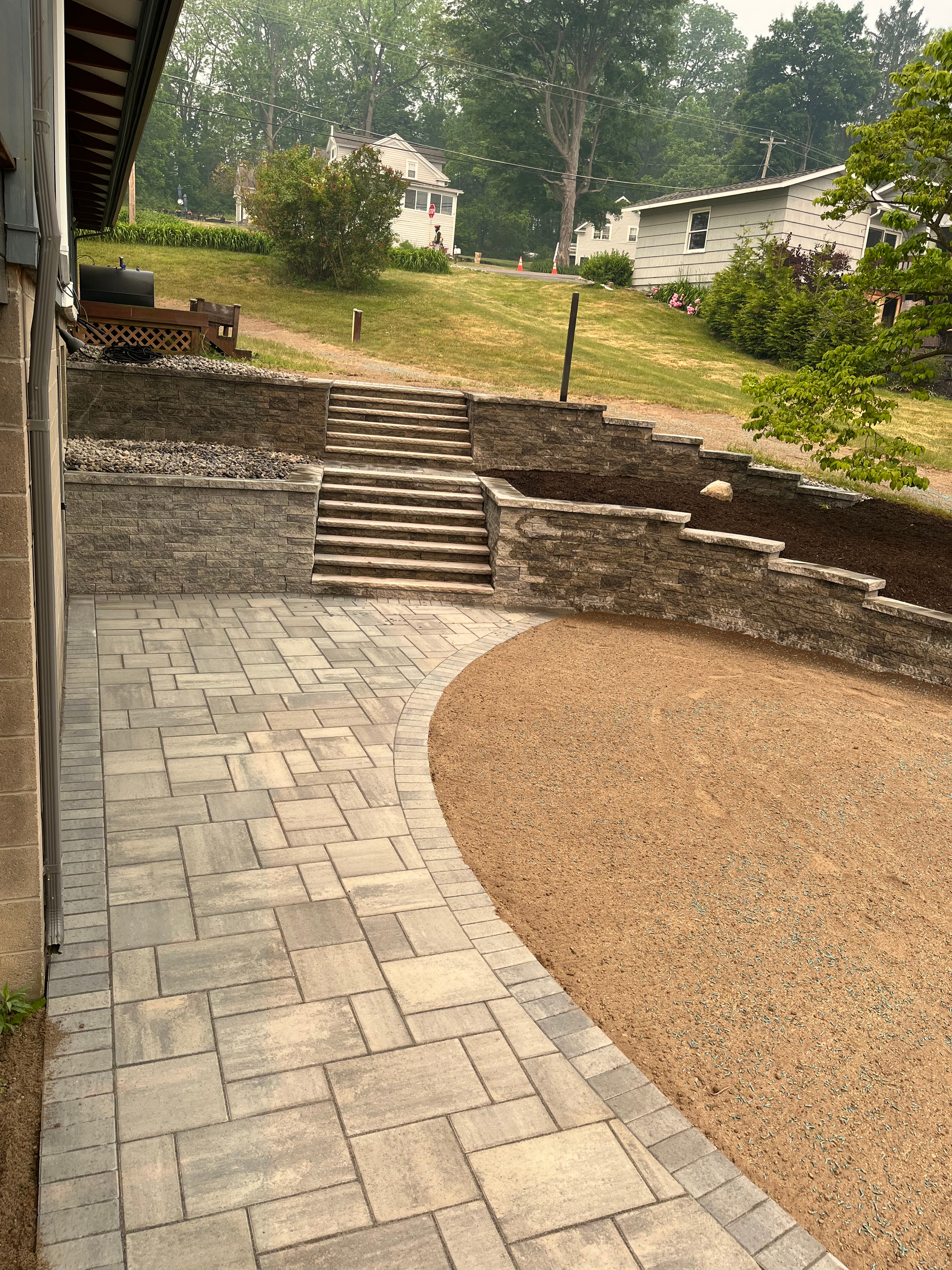 Newly paved patio with stone stairs leading up a landscaped backyard with grass, bushes, and trees.