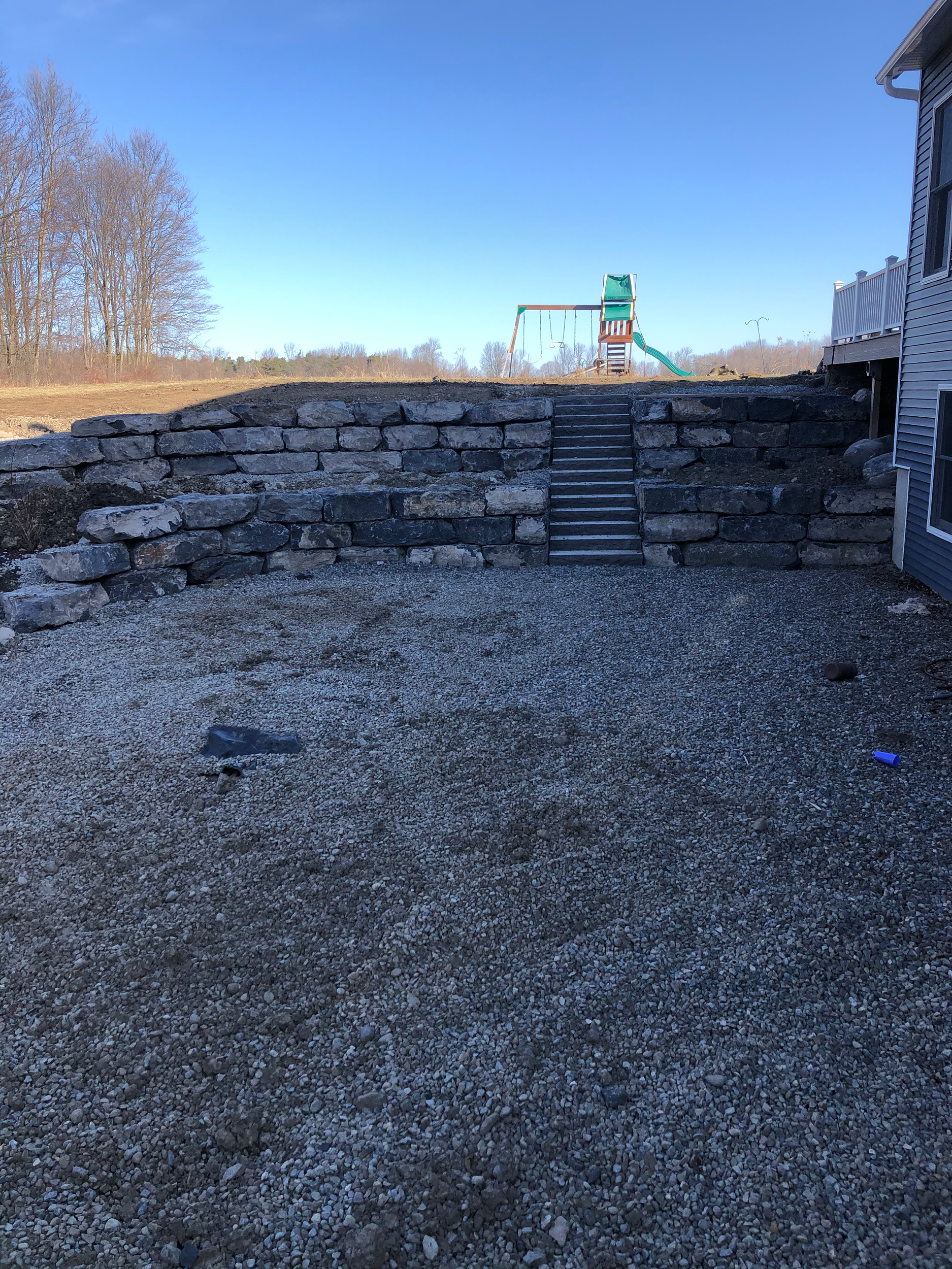 Backyard with stone retaining wall, gravel ground, and a playground with a slide and swings in the background under a clear blue sky.