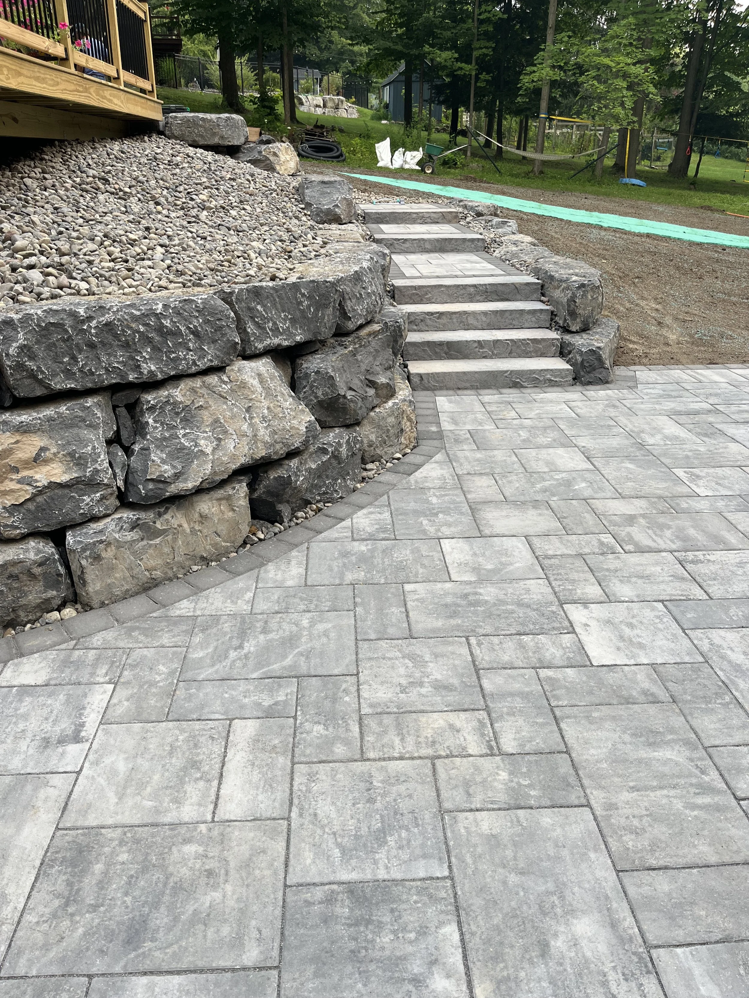 Outdoor stone patio with stairs leading up to a raised area, landscaped with large rocks and a rock retaining wall.