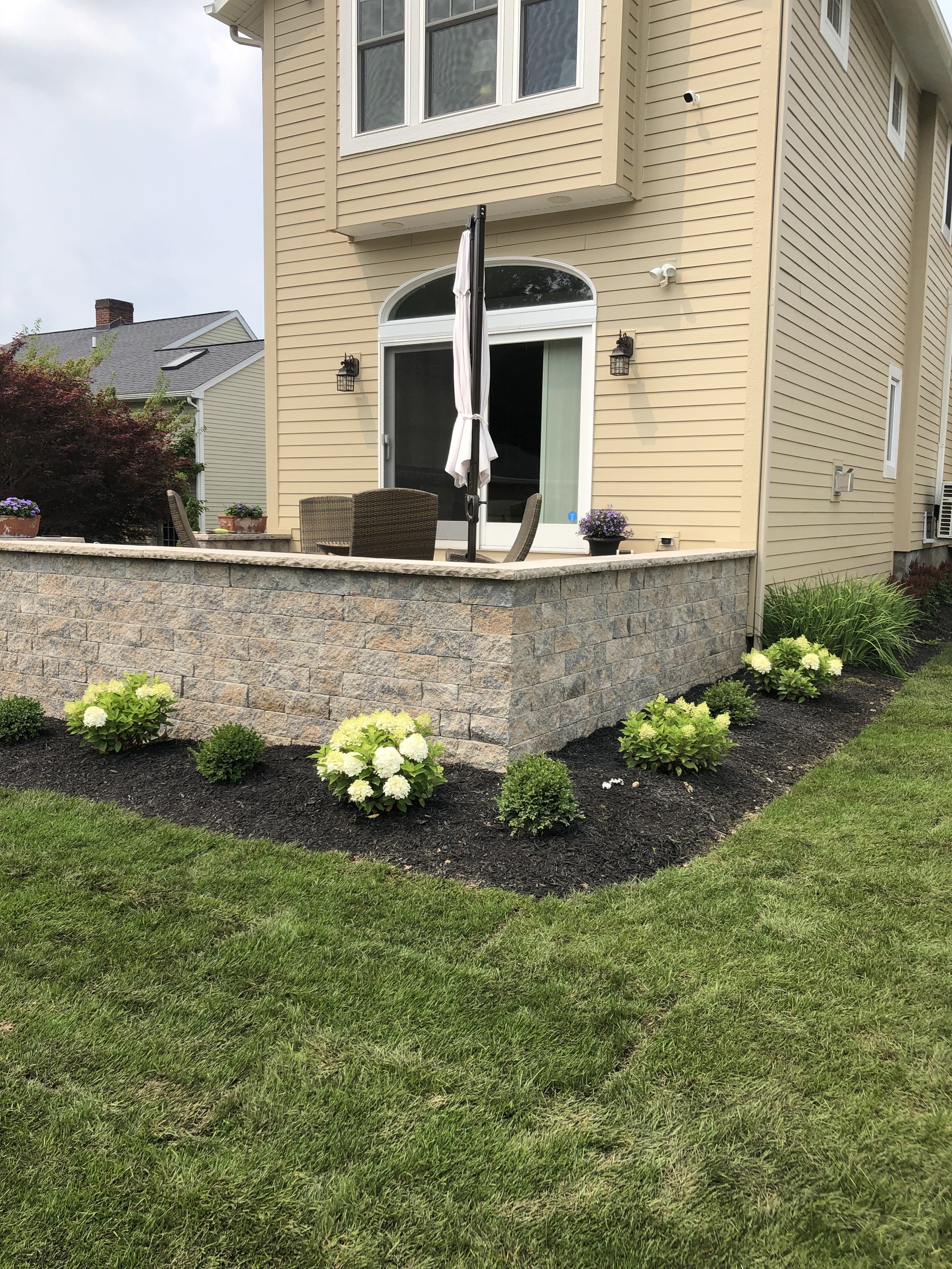 Backyard patio with outdoor furniture, potted plants, and a closed patio umbrella, surrounded by a garden with blooming plants and trimmed grass.