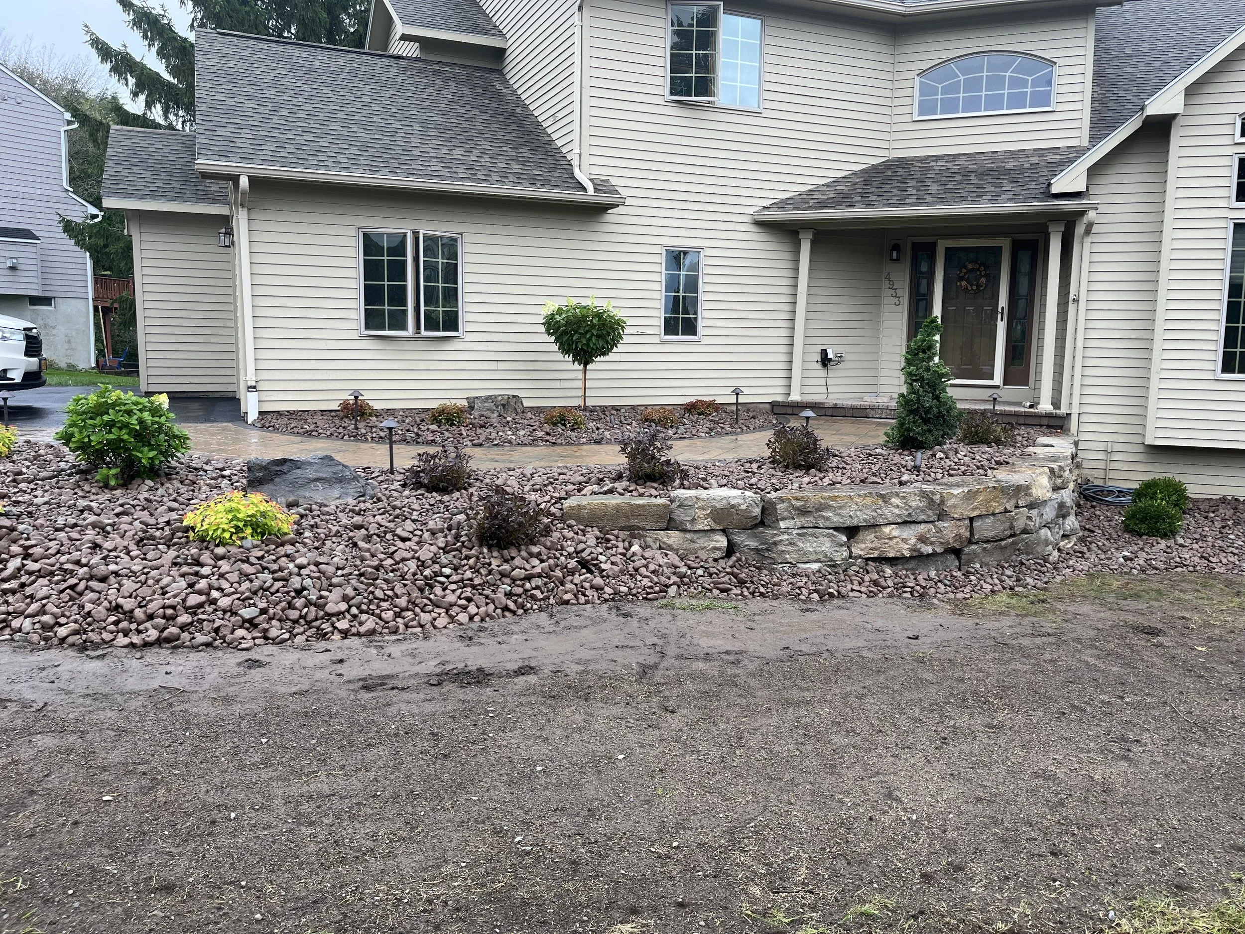 Front yard of a house with a landscaped garden featuring small bushes, a tree, rocks, and a stone retaining wall with a curved design, leading to a porch with a front door.