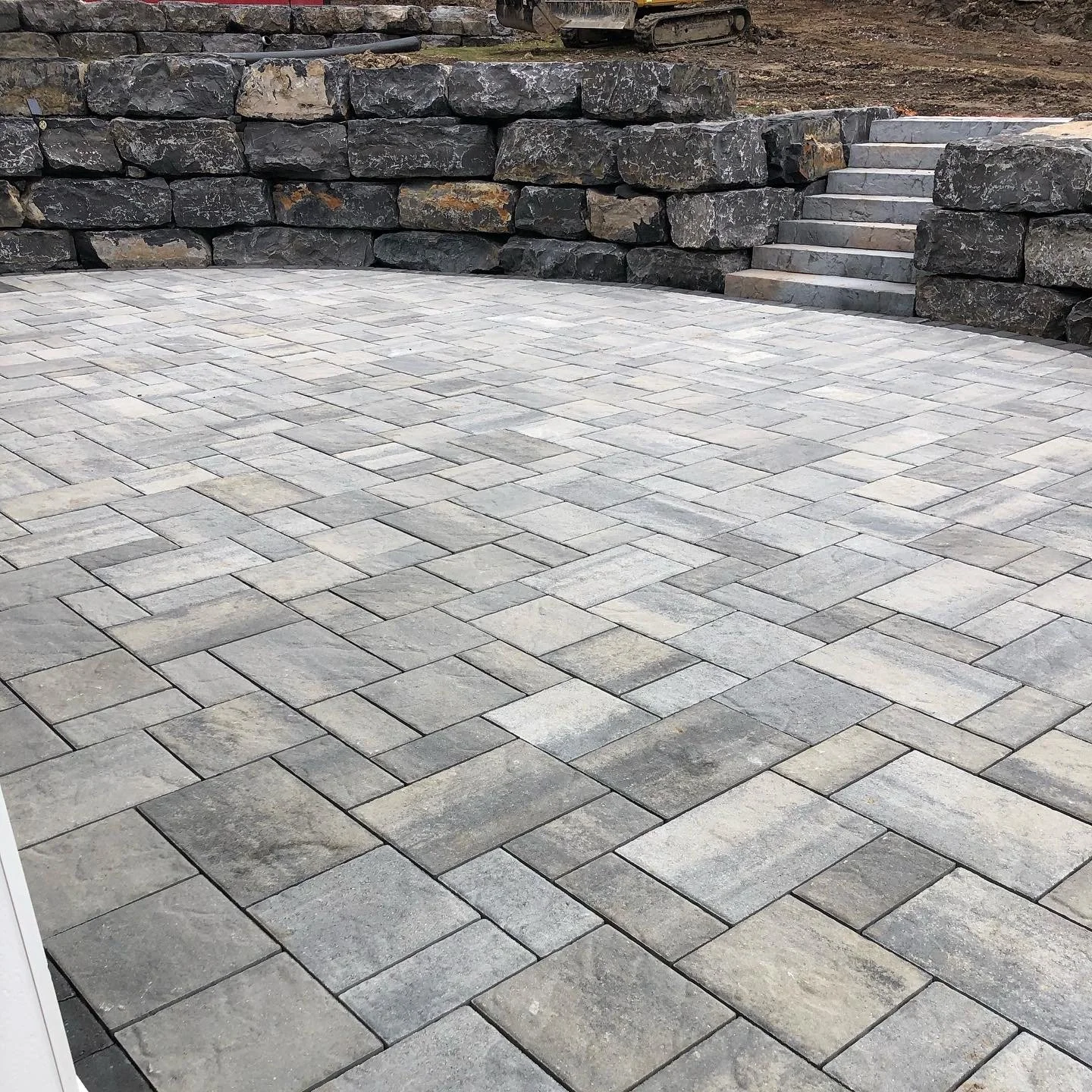 A stone patio with a retaining wall made of large dark rocks and a set of stone stairs.