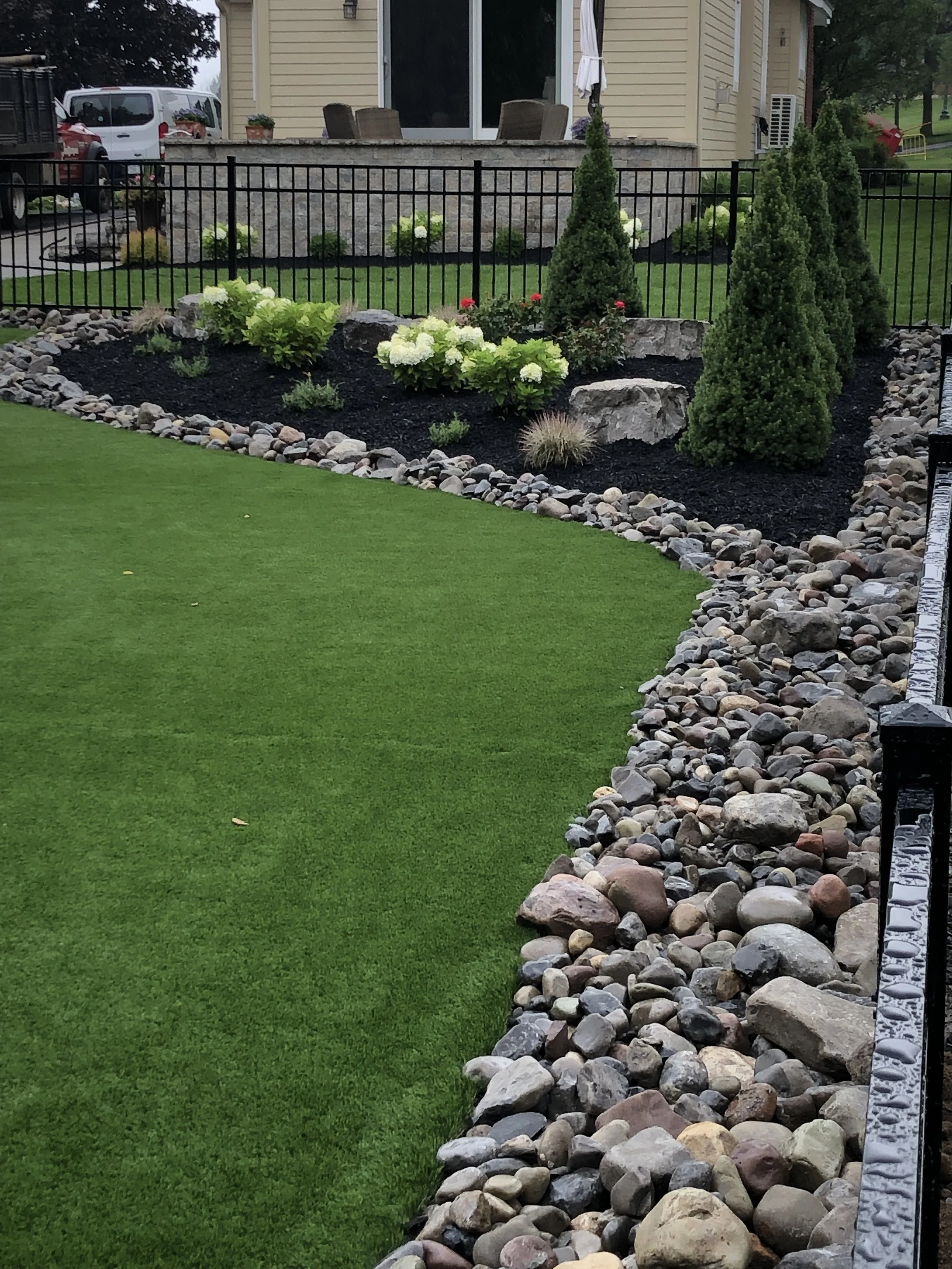 A landscaped backyard with a lush green lawn, a stone border, small shrubs, ornamental grasses, and three tall evergreen trees. There is a black metal fence and a patio area with chairs and an umbrella in the background.