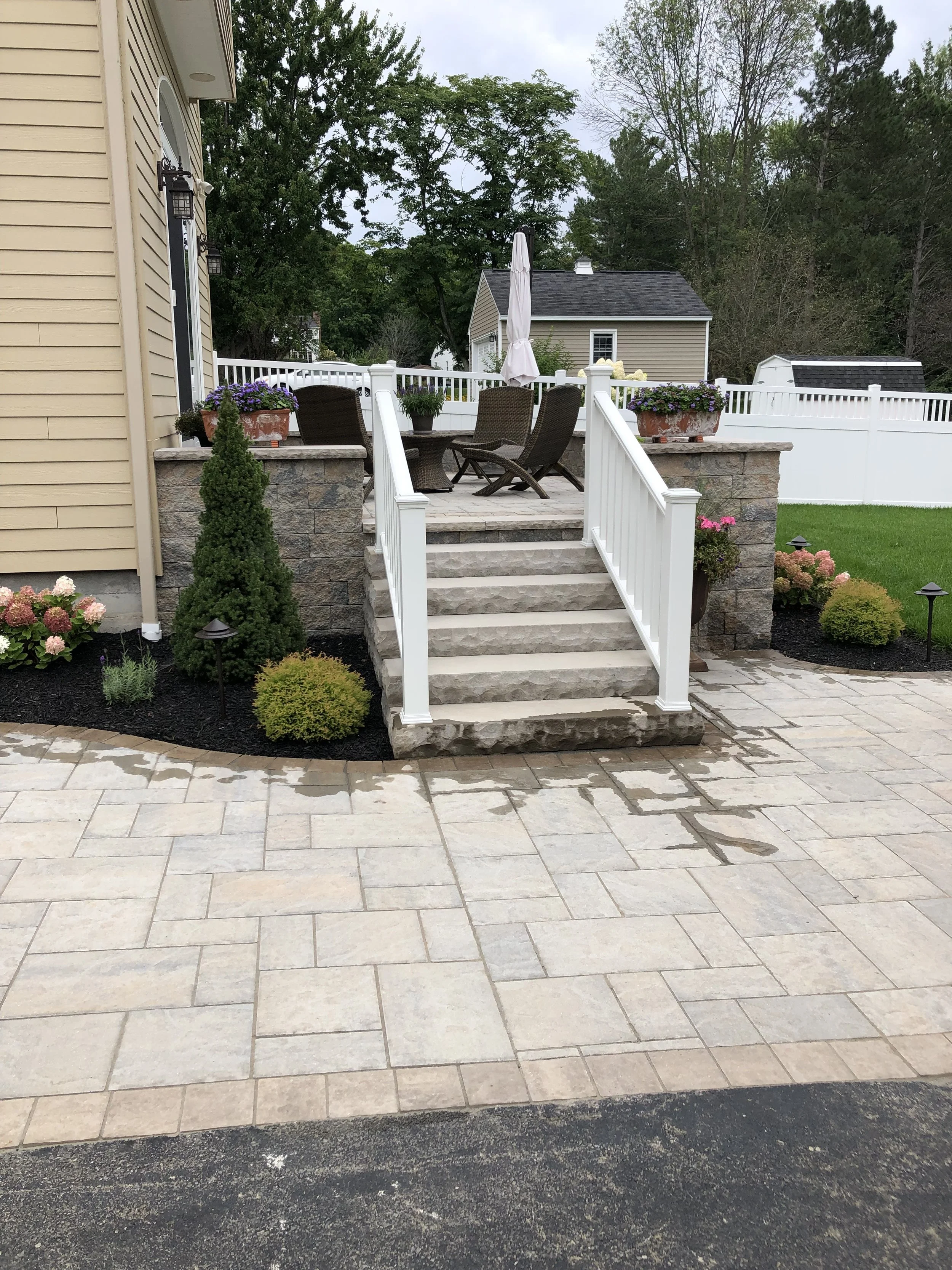 A backyard patio area with a stone staircase leading up to a raised deck with outdoor chairs and a table under an umbrella. The deck is surrounded by a white railing and decorated with potted flowers and shrubs. There is a house on the left side and 