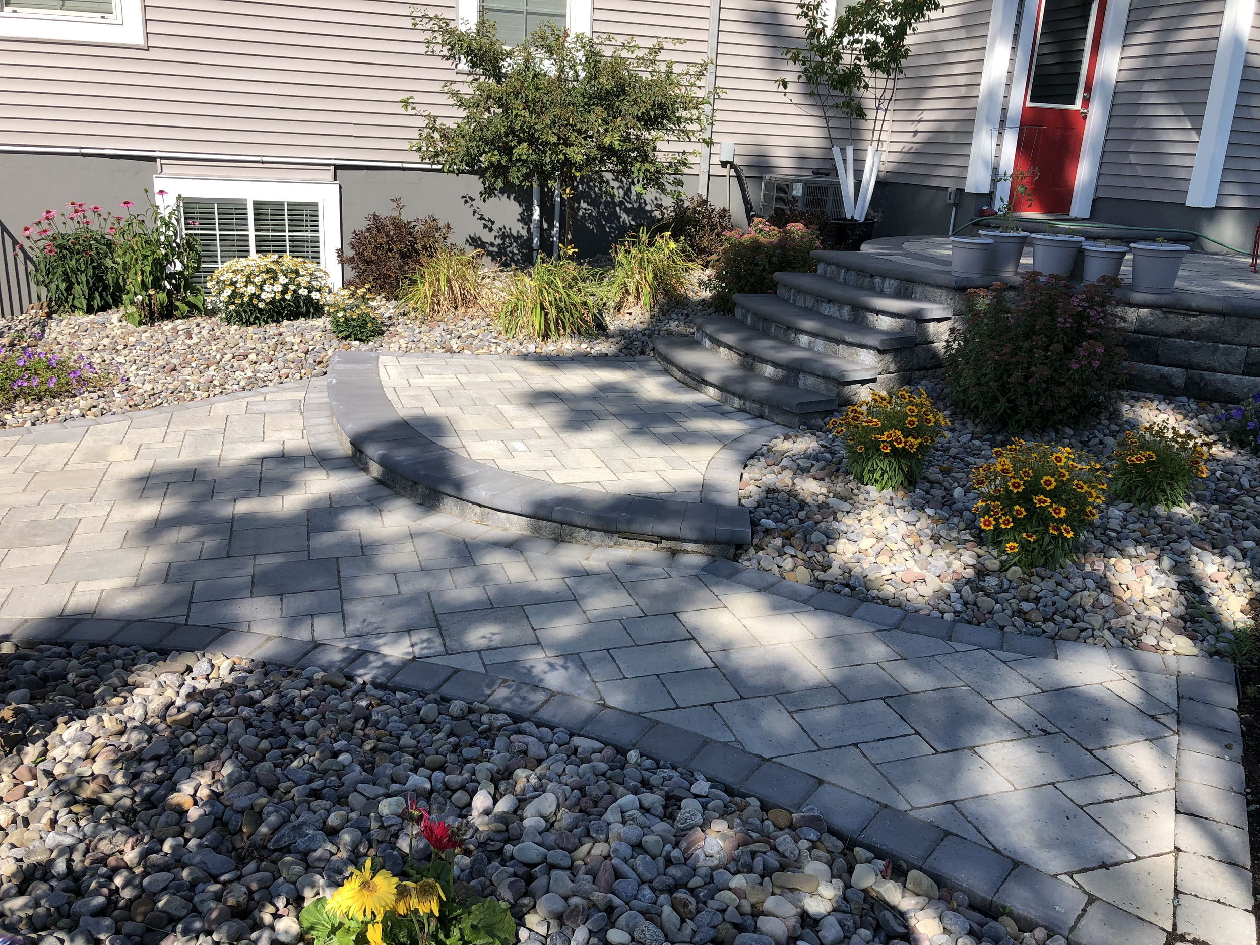 A backyard landscaped with a stone pathway, steps, and flower beds with colorful flowers including yellow, purple, and pink blossoms. There are potted plants on the upper patio, a bush, and a tree with green leaves, with a house wall in the backgroun