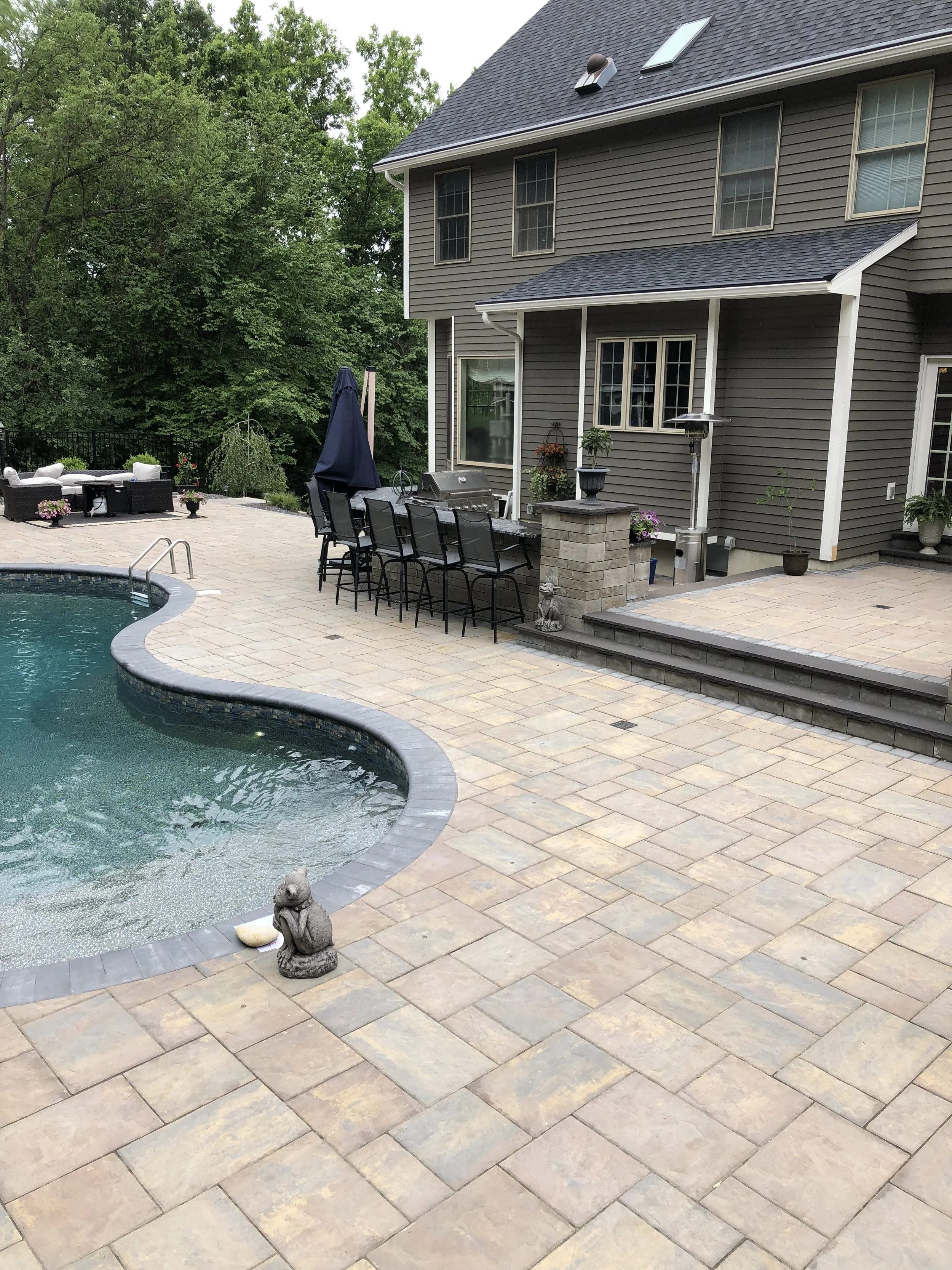 Backyard patio with a swimming pool, outdoor seating area, a barbecue grill, potted plants, and a house with gray siding and multiple windows, surrounded by green trees.
