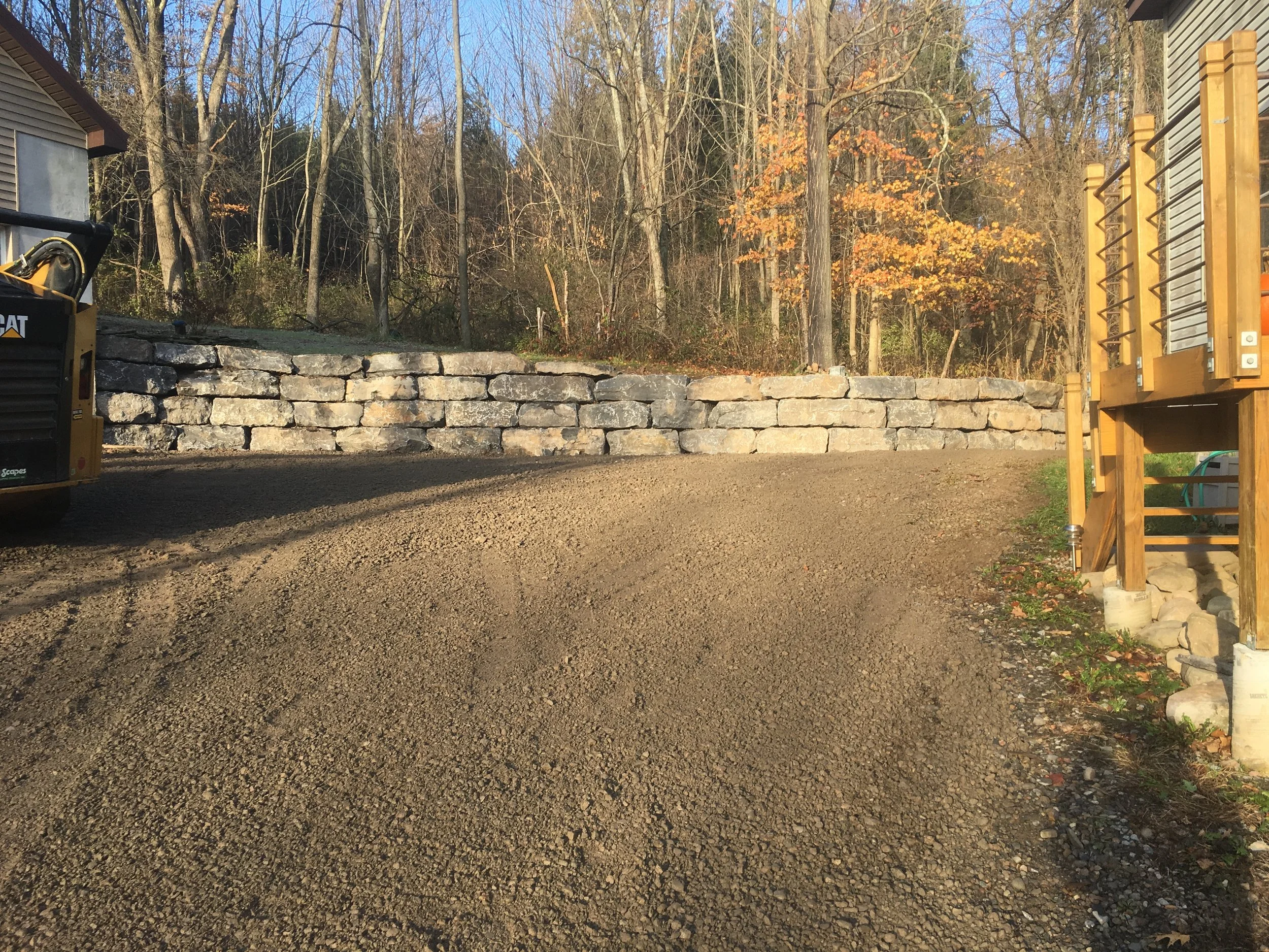A gravel driveway or path bordered by a stone retaining wall and a wooden deck, with trees in the background showing autumn foliage.