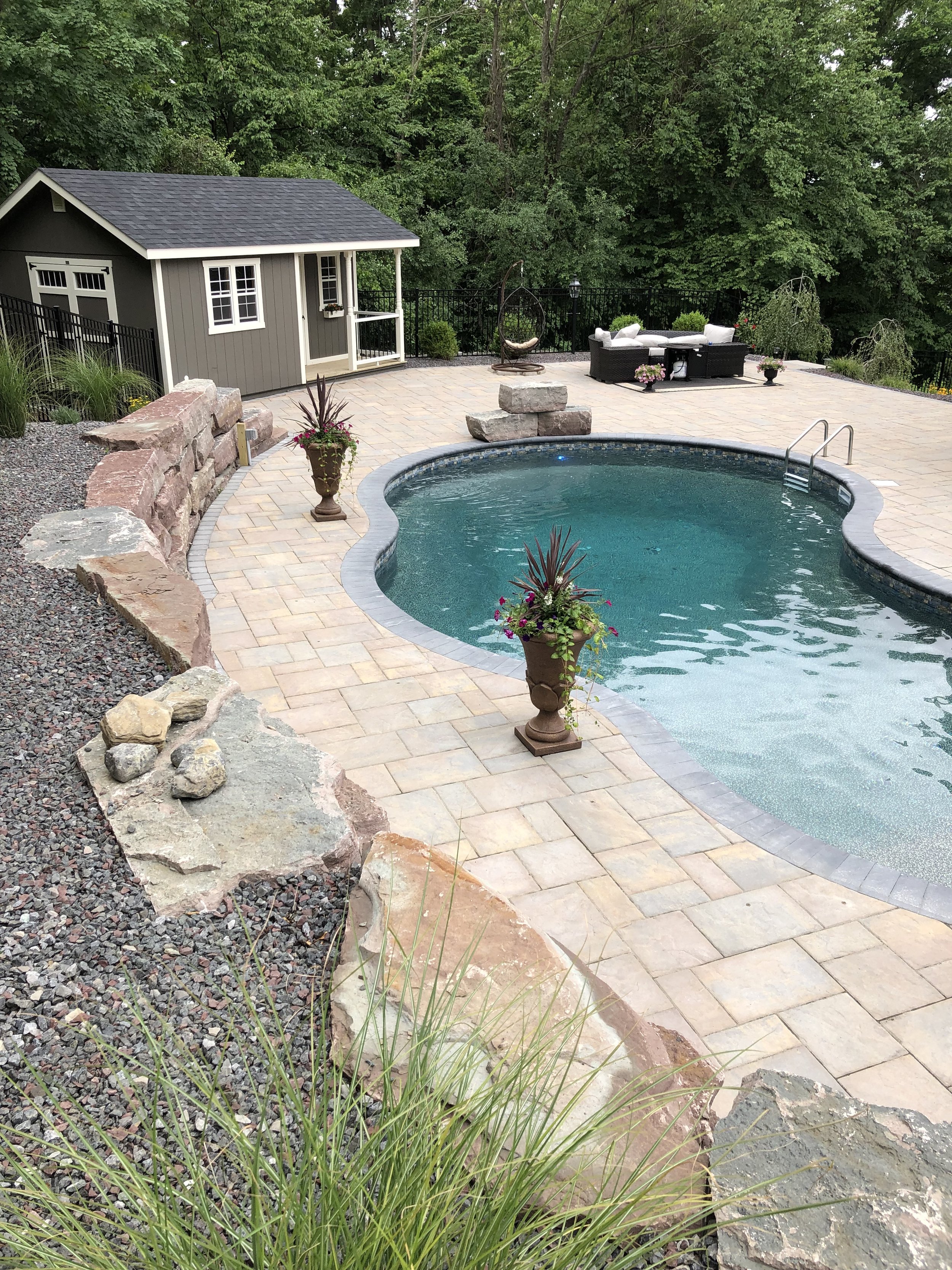 Backyard swimming pool with stone border, surrounded by potted plants, a small shed, and outdoor seating area, with green trees in the background.