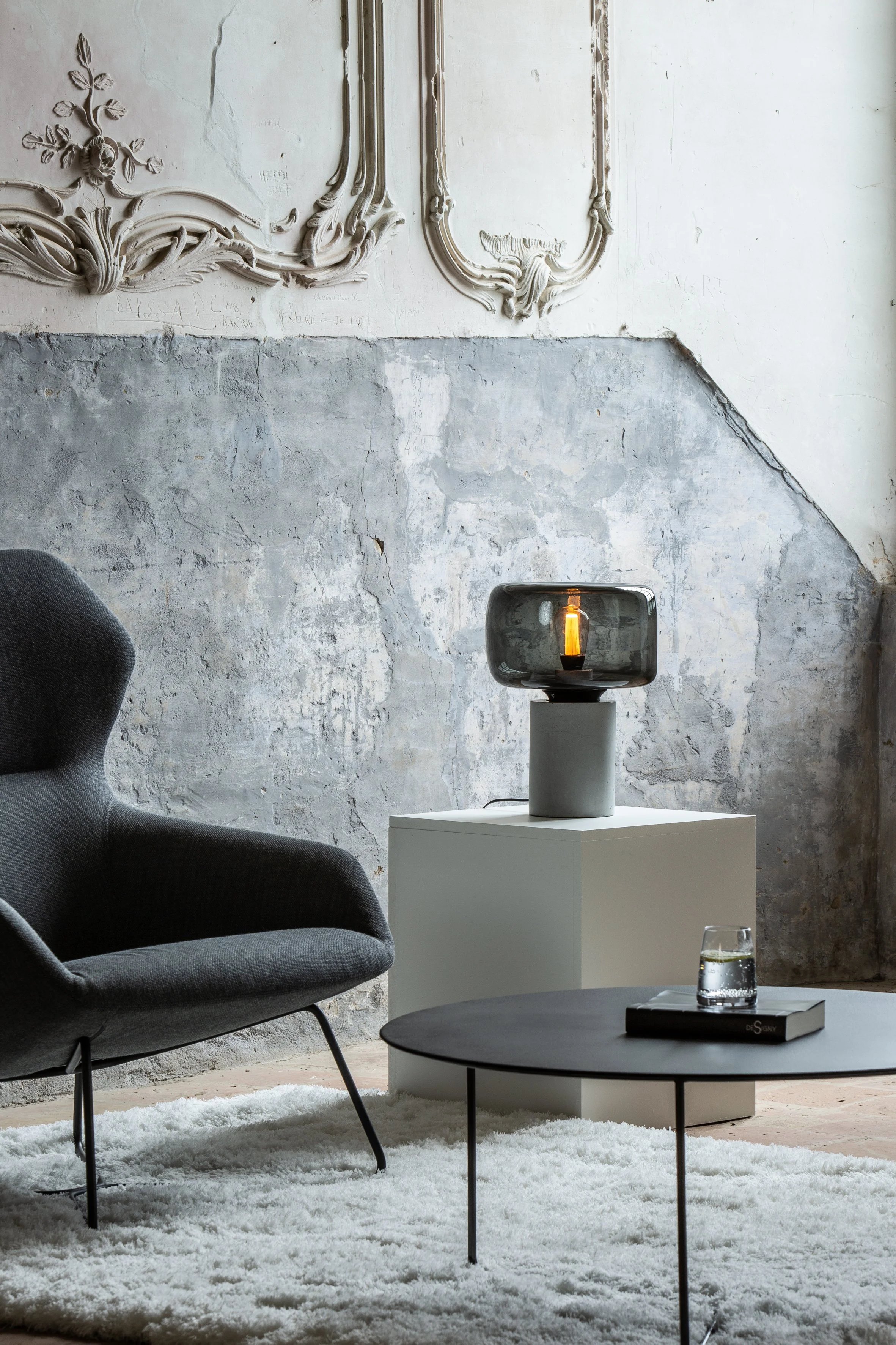 Salon moderne avec un fauteuil gris, une table basse ronde noire, un tapis blanc, une lampe sur un socle blanc, un mur en pierre et des décorations murales ornementales.