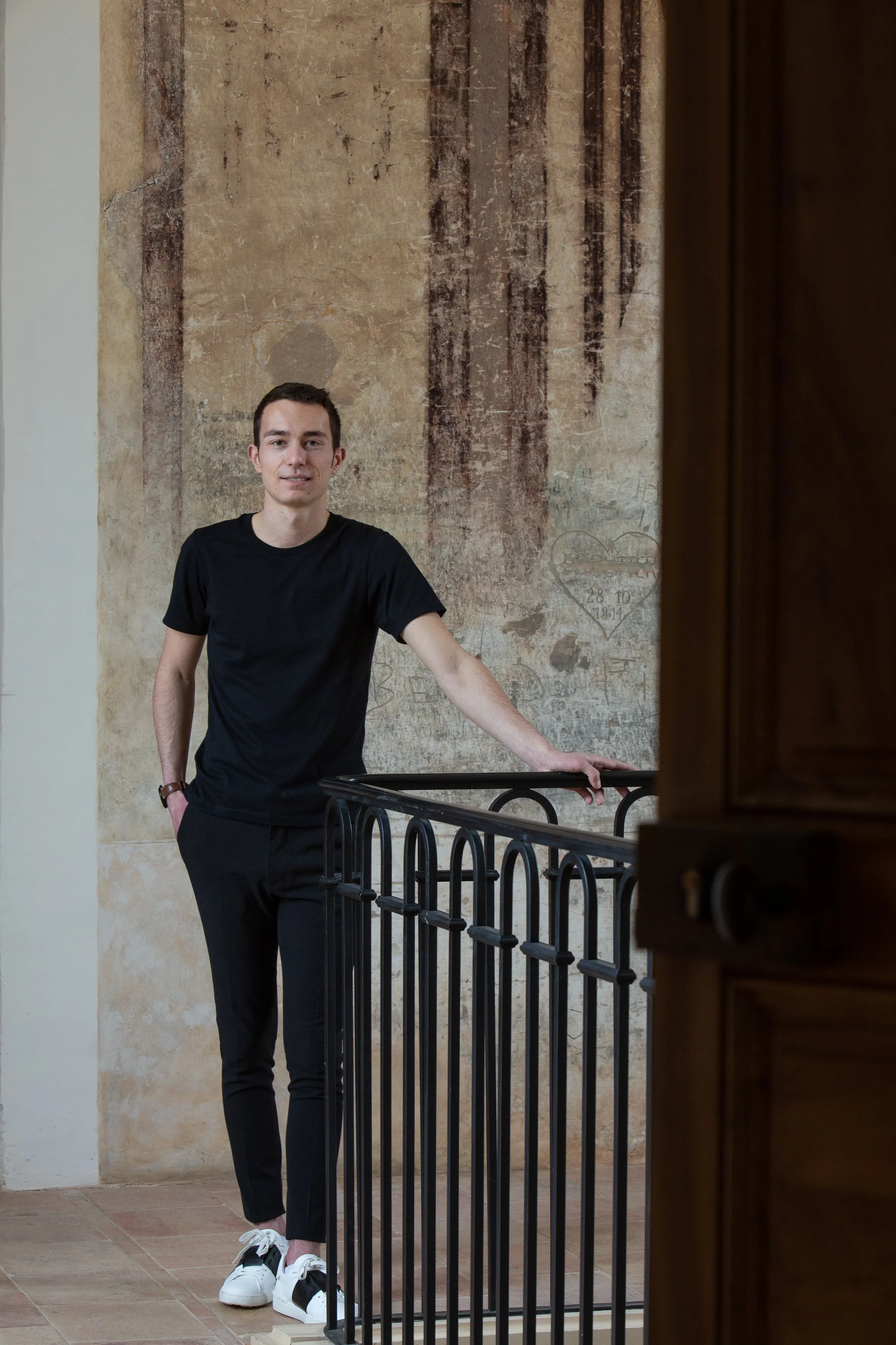 Un jeune homme en t-shirt noir et pantalon noir se tient debout près d'une rampe en métal dans une pièce avec un mur en pierre ancienne.