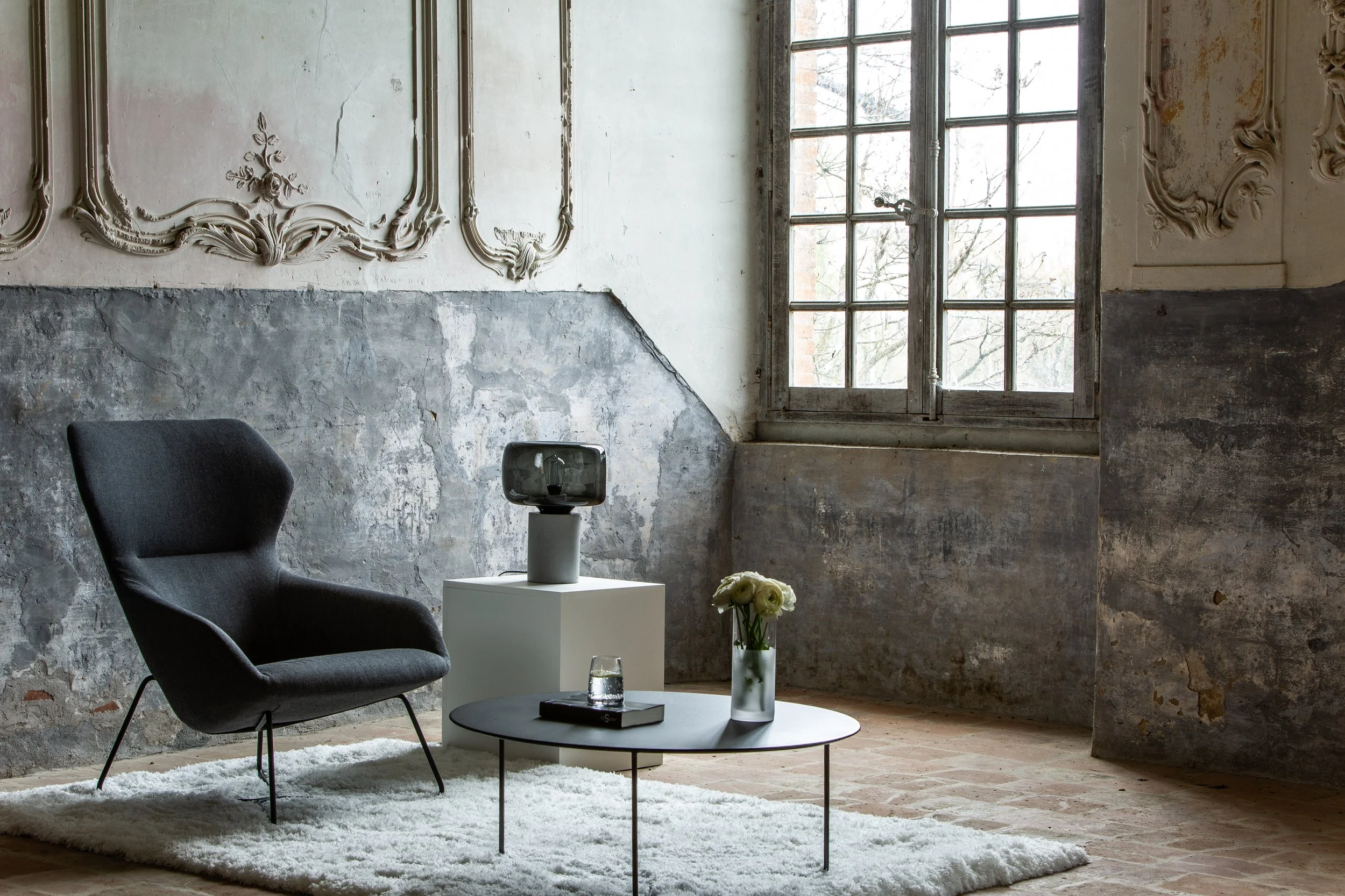 Intérieur avec un fauteuil noir, une table basse ronde avec un vase à fleurs blanc, un livre et un verre d'eau, une lampe sur un socle blanc, et une grande fenêtre.