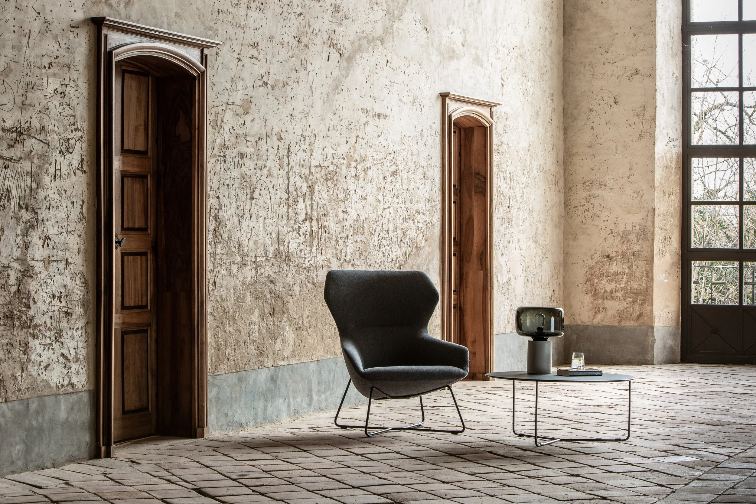 Intérieur d'une pièce avec murs usés, deux portes en bois, un fauteuil noir, une table basse avec une enceinte et une tasse, grande fenêtre laissant entrer la lumière naturelle.