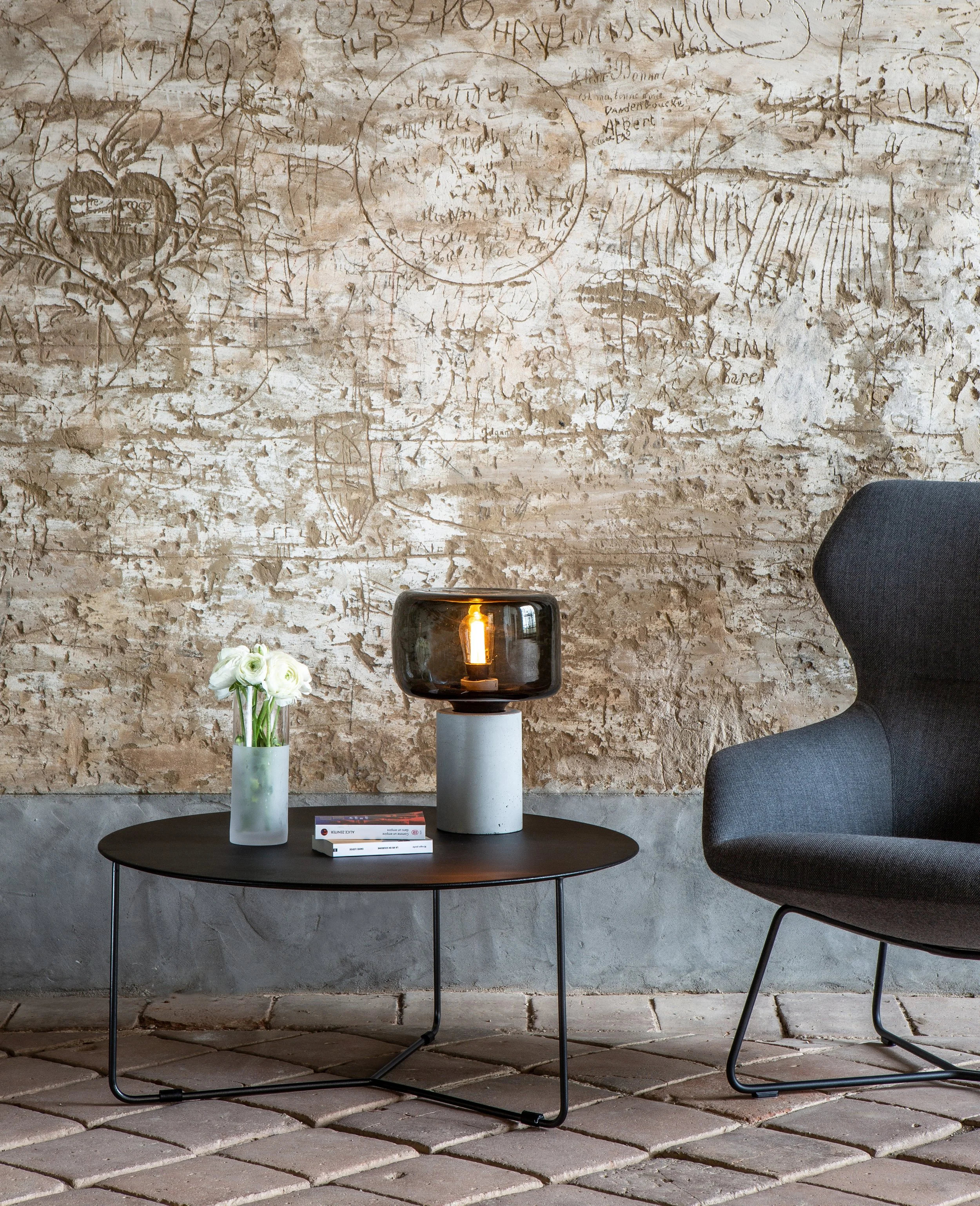 Un salon moderne avec une table basse noire, une lampe en verre noire, un vase avec des fleurs blanches, un fauteuil gris foncé et un mur en pierre décoré d'esquisses et de dessins.