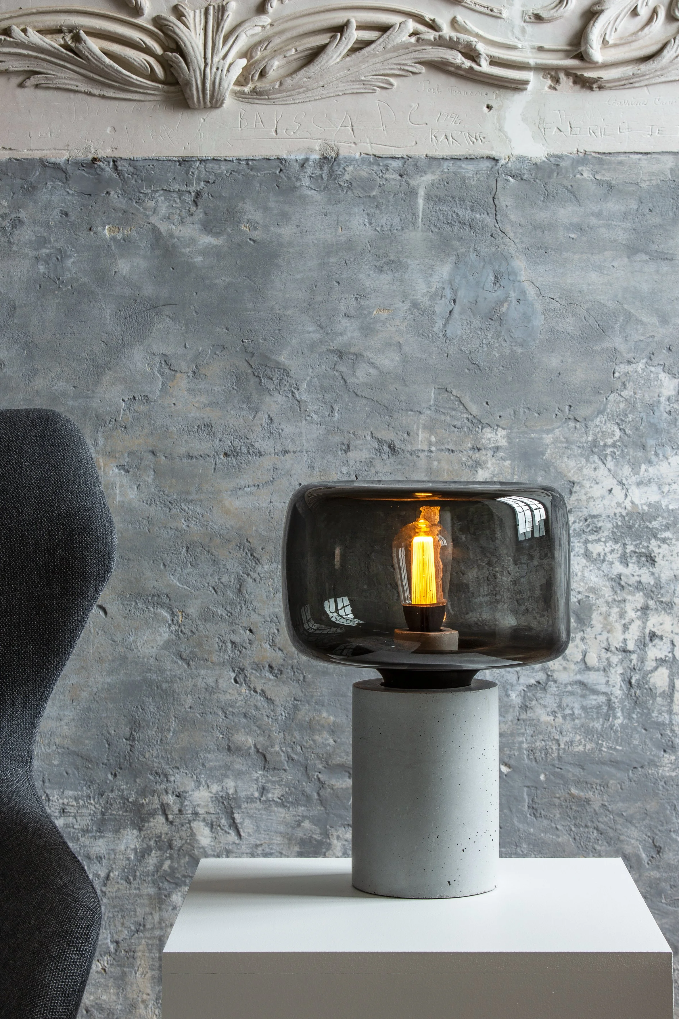 Une lampe moderne avec un abajour en verre foncé, posée sur une base en béton, dans un intérieur avec un mur en béton brut.