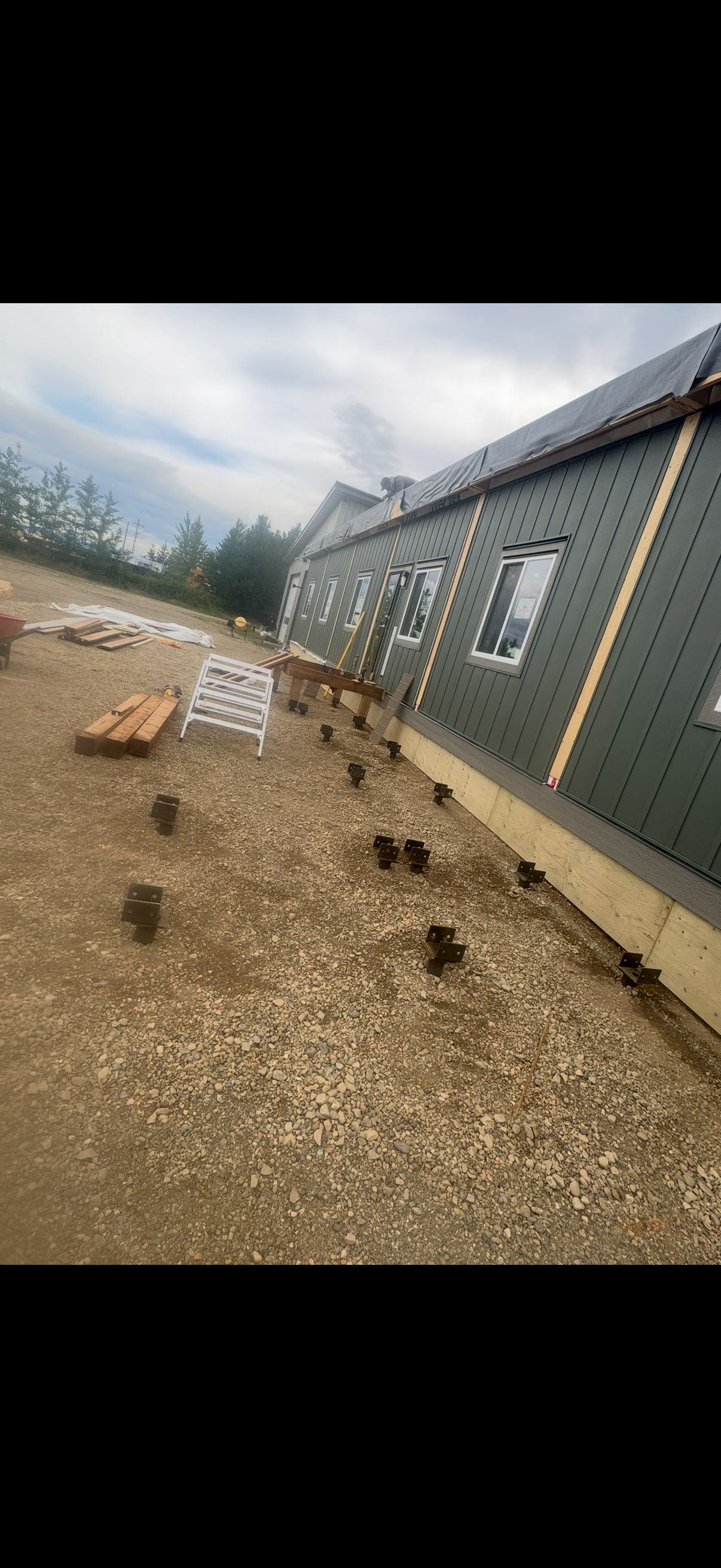 Construction site with a house being built, showing metal siding and multiple windows, with foundation piers and building materials on the ground.