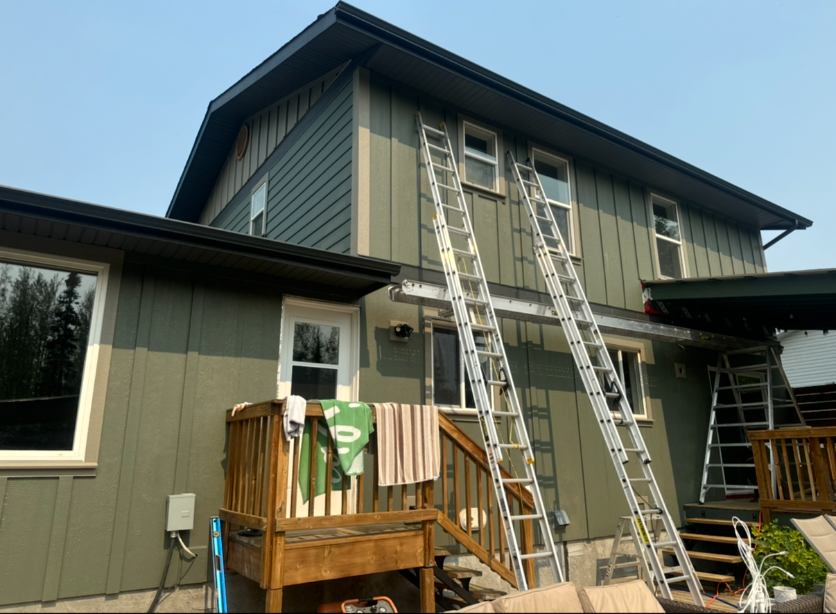 A two-story house with dark green vertical siding, multiple windows, and a metal roof. Ladders are set against the house, indicating ongoing exterior work. There are towels and a towel hanging on the wooden deck railing in the backyard.