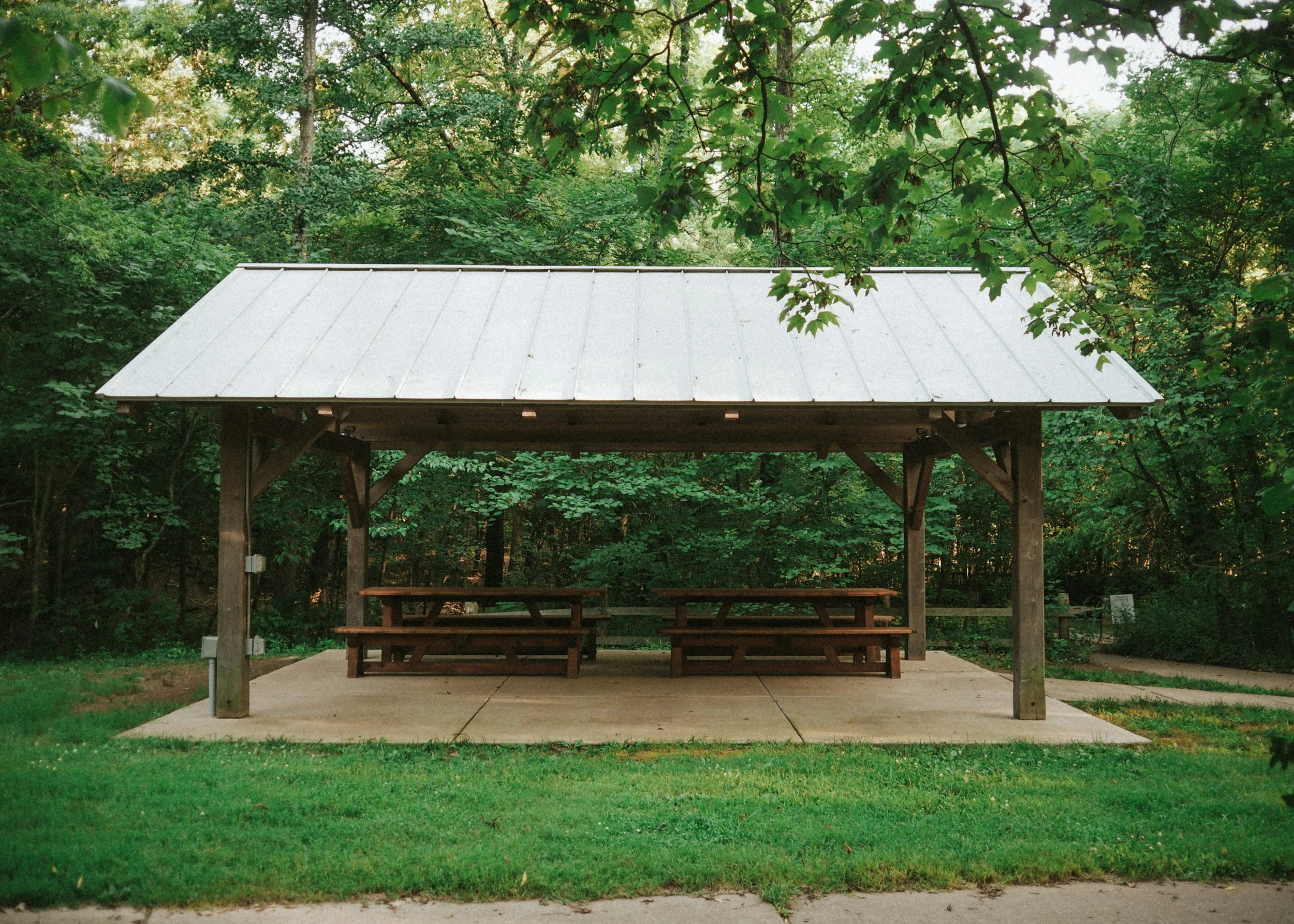 Wooden pavilion with picnic tables on a concrete slab, surrounded by green trees and grass.