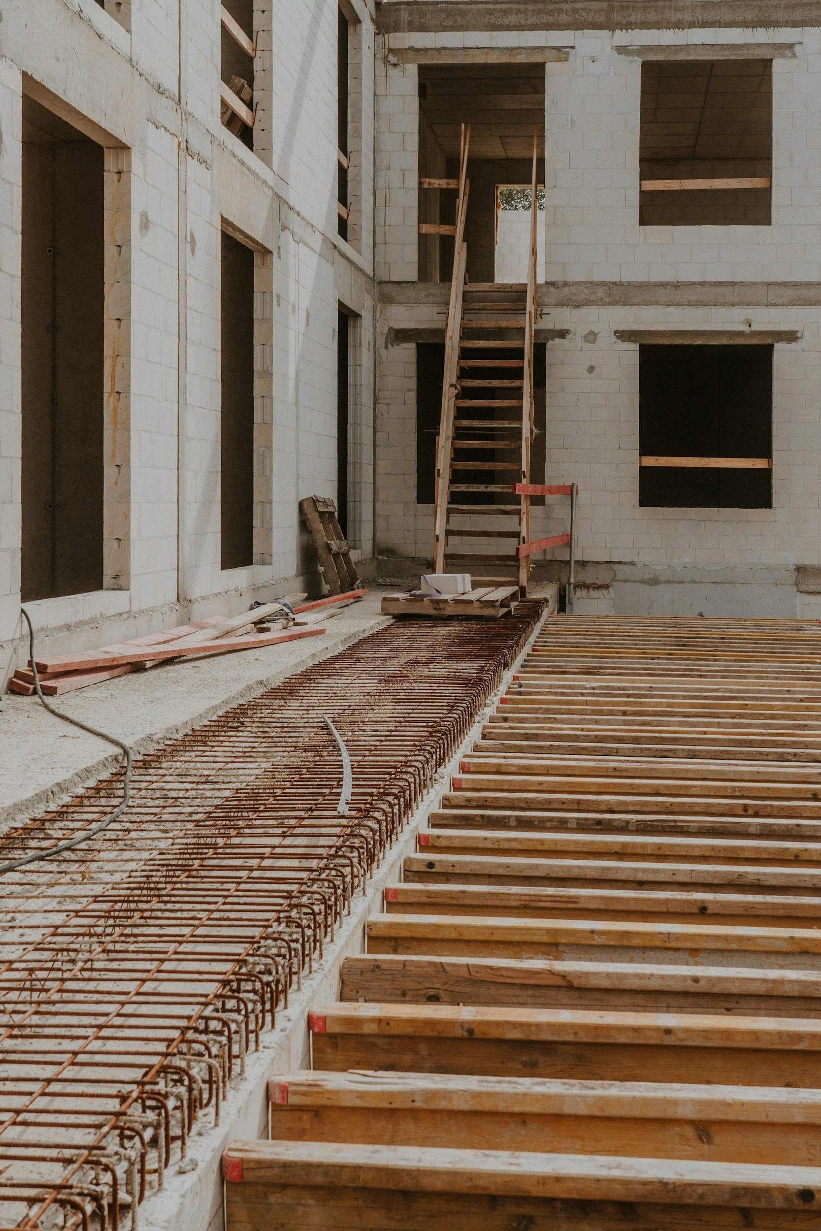 Interior of a building under construction with exposed brick walls, wooden floor framing, metal rebar, and a wooden staircase.