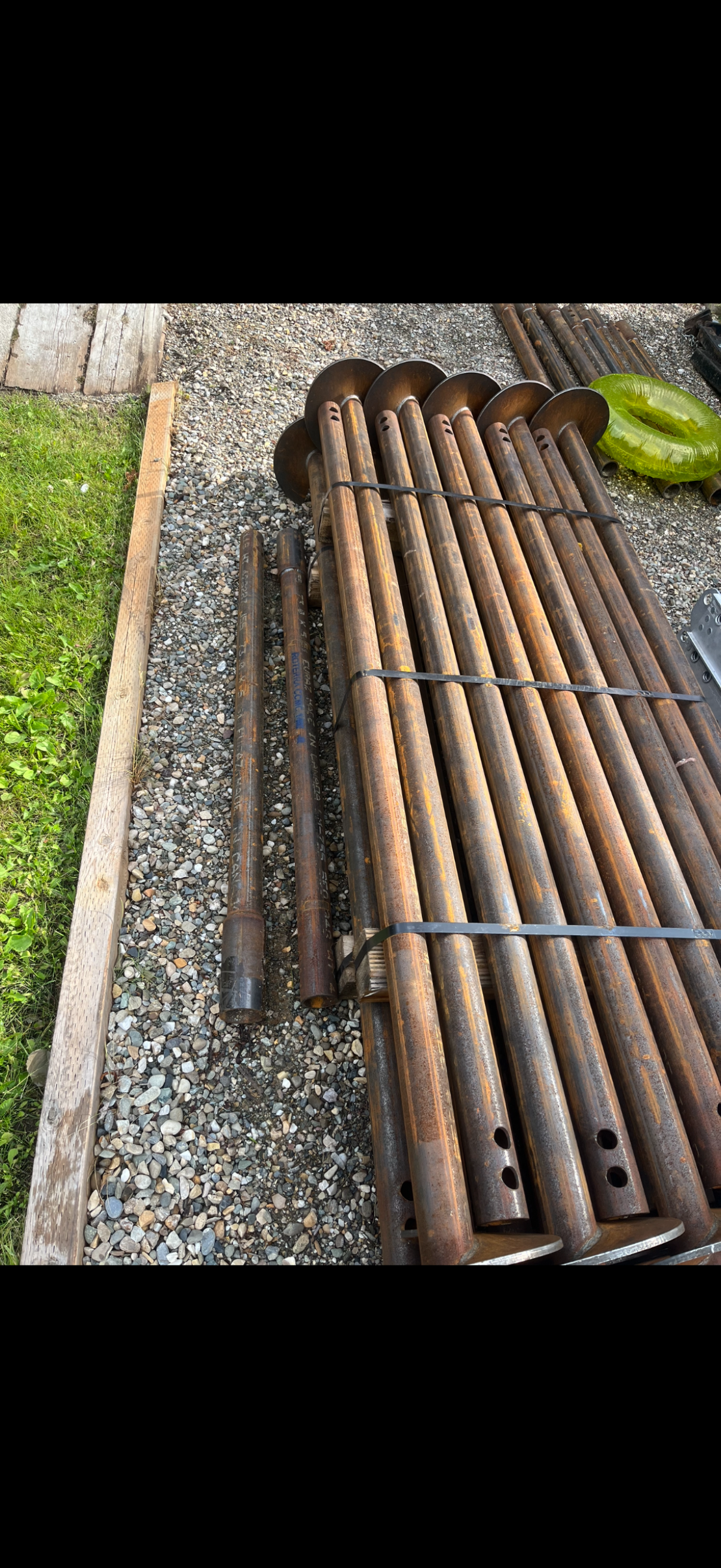 A pile of rusty steel pipes and metal parts stacked outside on gravel next to grass, secured with metal straps.