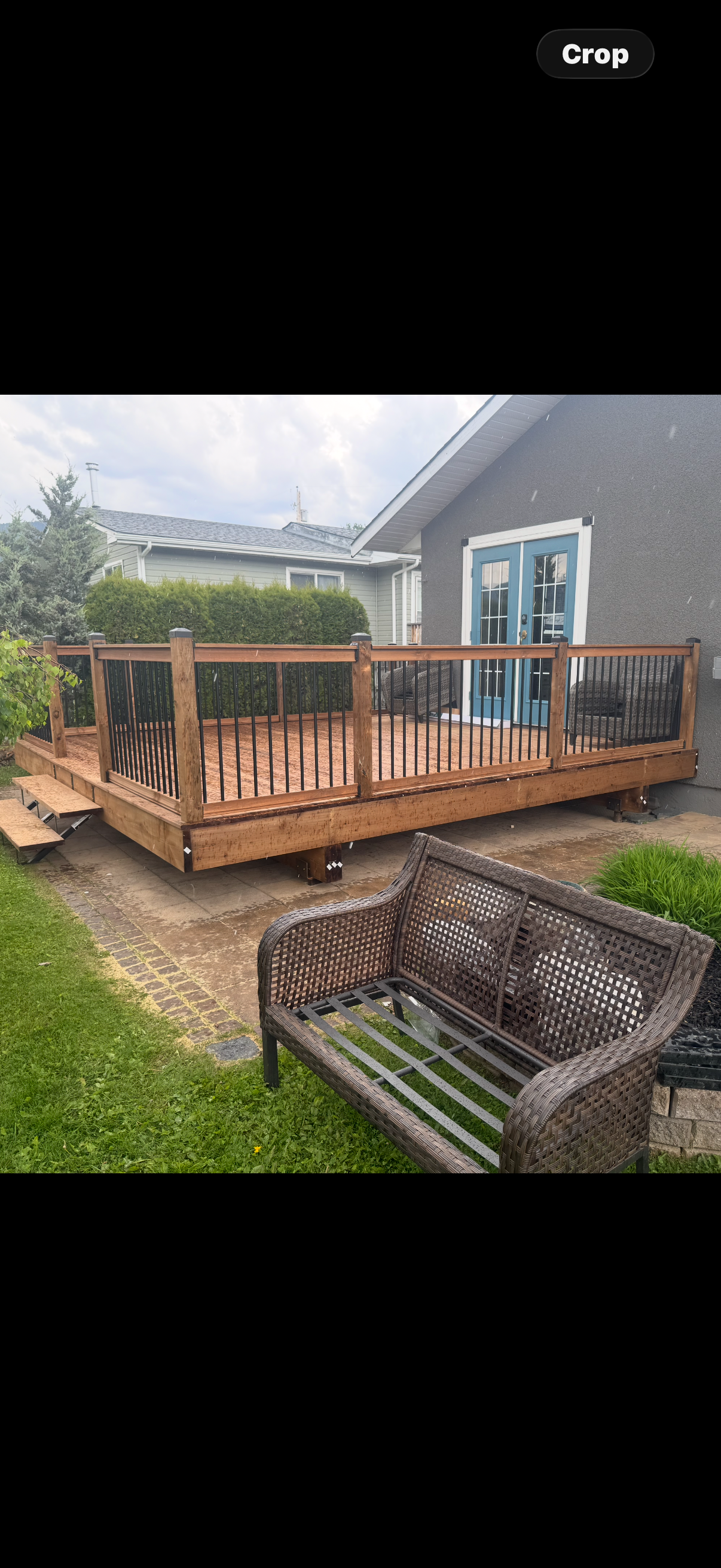 Newly built wooden deck with black metal railing attached to the back of a house with blue French doors, a wicker bench in the yard, and a neighboring house with light green siding in the background.