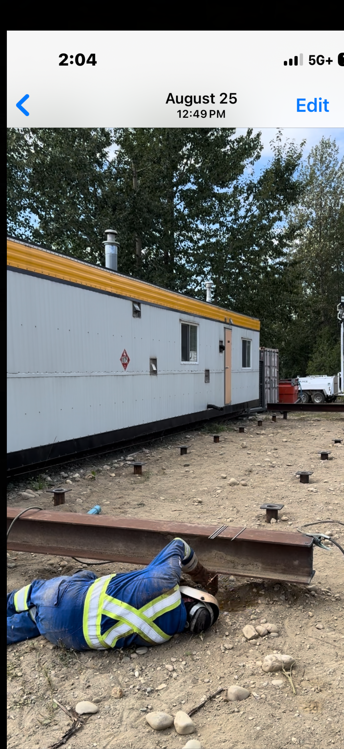 A construction worker wearing a blue safety vest and helmet working on a steel beam on the ground outside near a building with a yellow roof trim. The area is rocky with some wood and construction materials nearby. Trees are in the background.