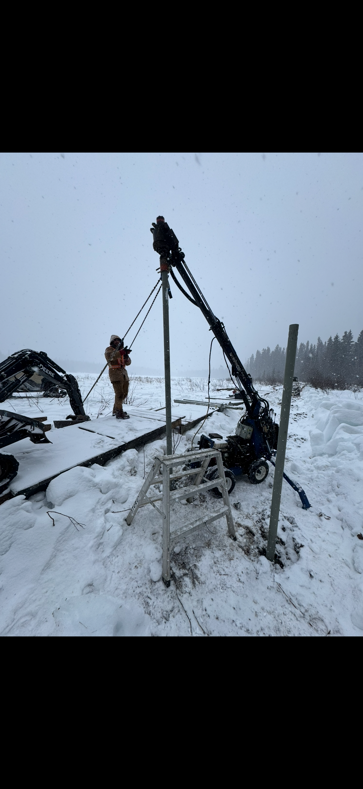 Two workers in snow using a drill to install a utility pole, with snow-covered ground, winter landscape, and equipment surrounding them.