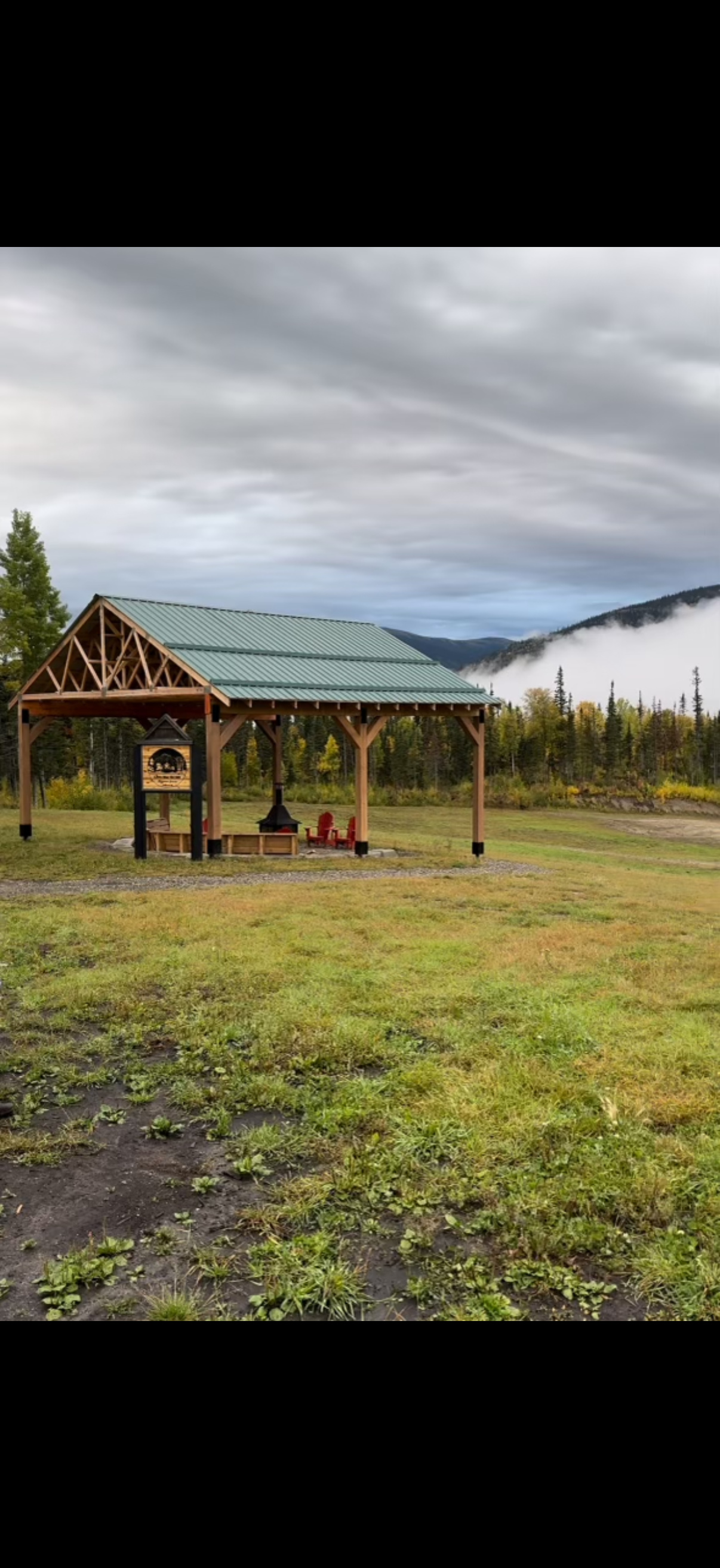 A wooden shelter with a green metal roof, located in a grassy area with mountains and pine trees in the background, under a cloudy sky.