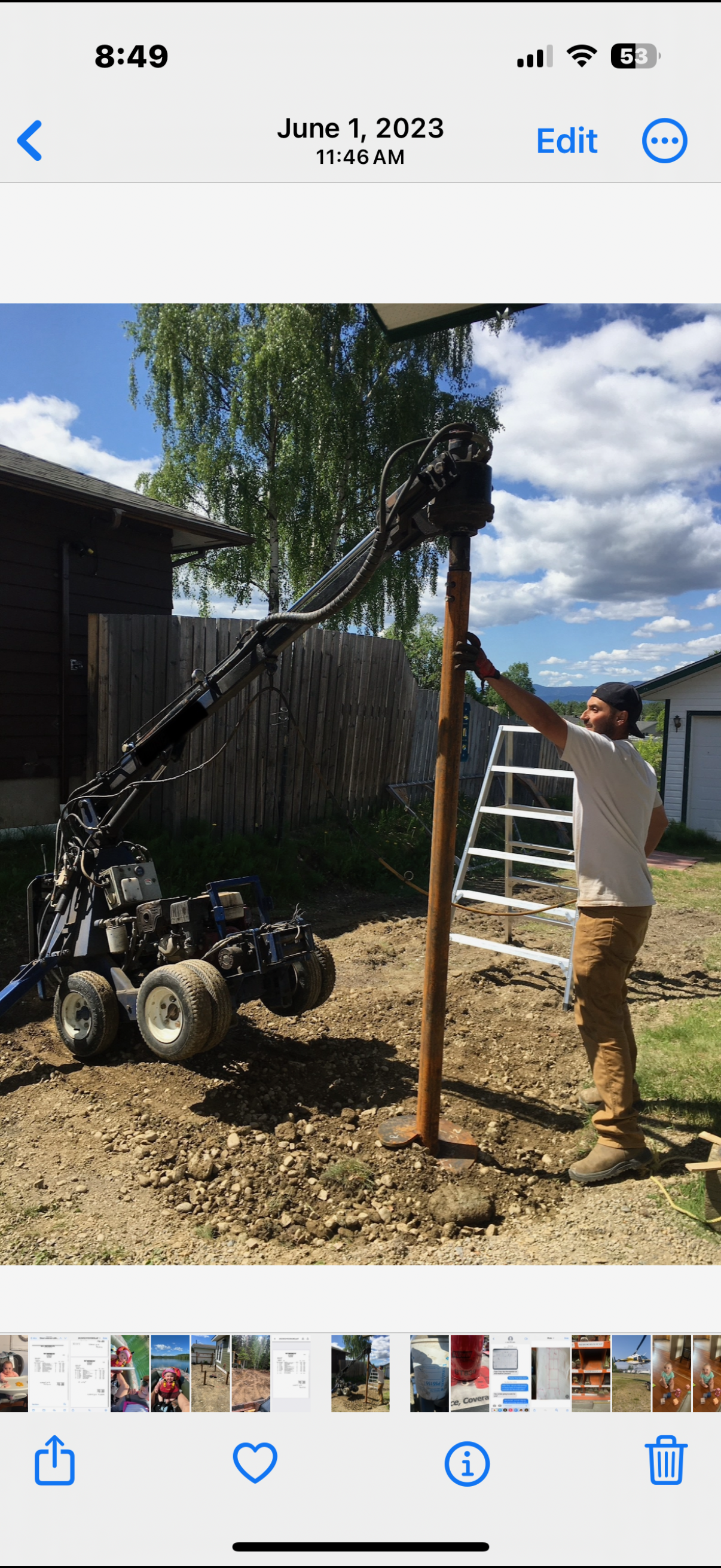 A man in a white shirt and khaki pants operating a small drilling rig outdoors. The rig is attached to a rusty metal post, and the man is holding the post steady. The background includes a ladder, a house, a wooden fence, trees, and a partly cloudy s