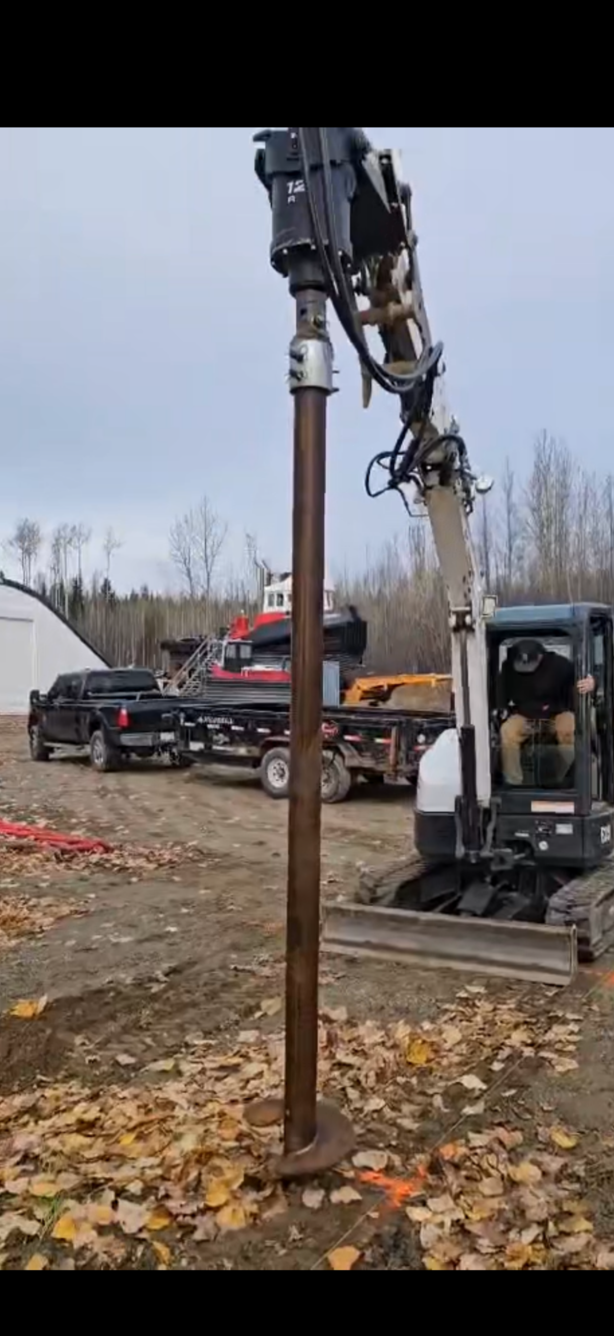 A construction site with a small excavator drilling a hole into the ground near a large metal pipe, surrounded by fallen leaves, with trucks in the background and a cloudy sky.