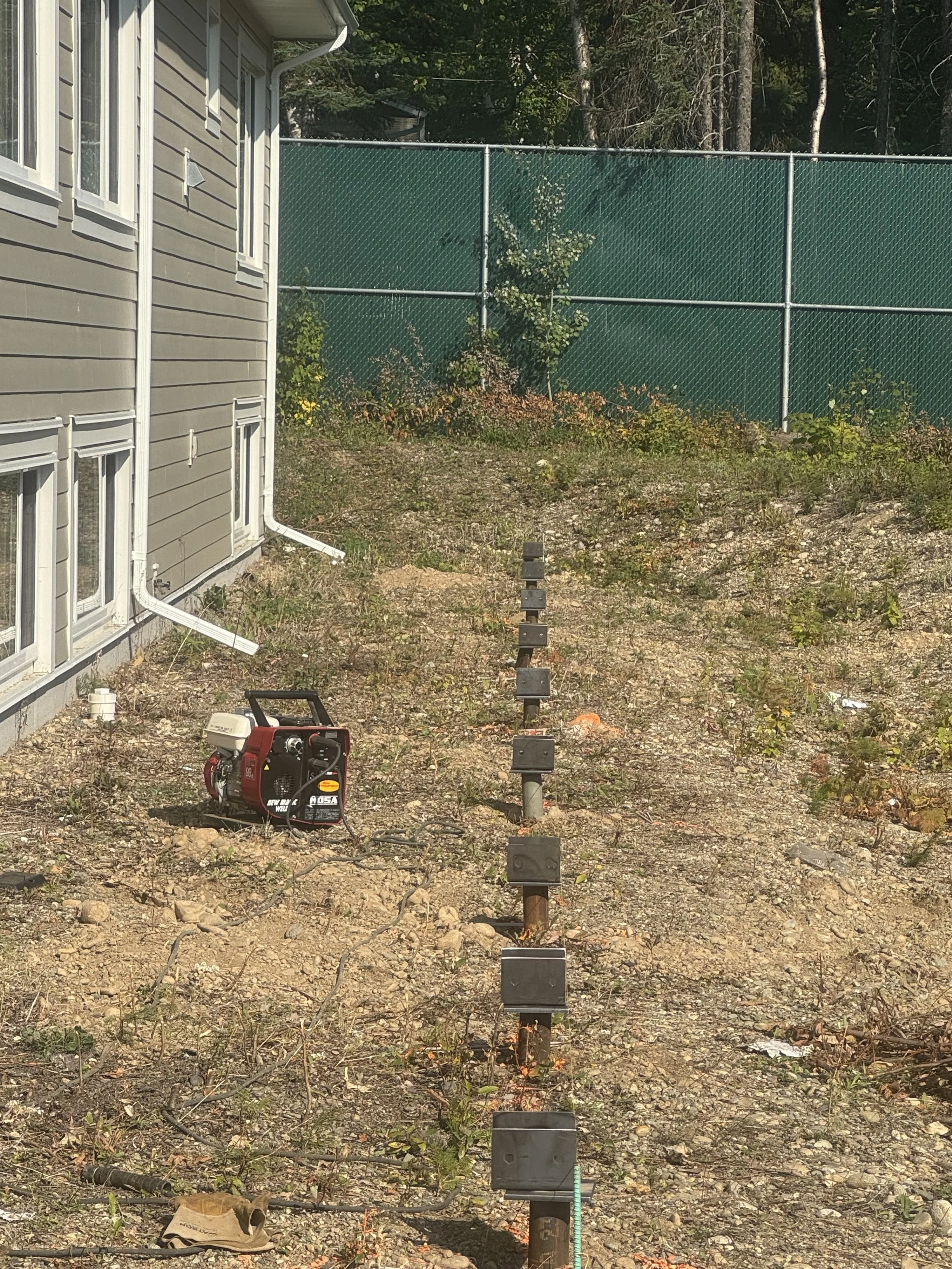 Construction site adjacent to a house with a green fence, electric generator, and metal posts in the ground for future installation of fencing or support structures.