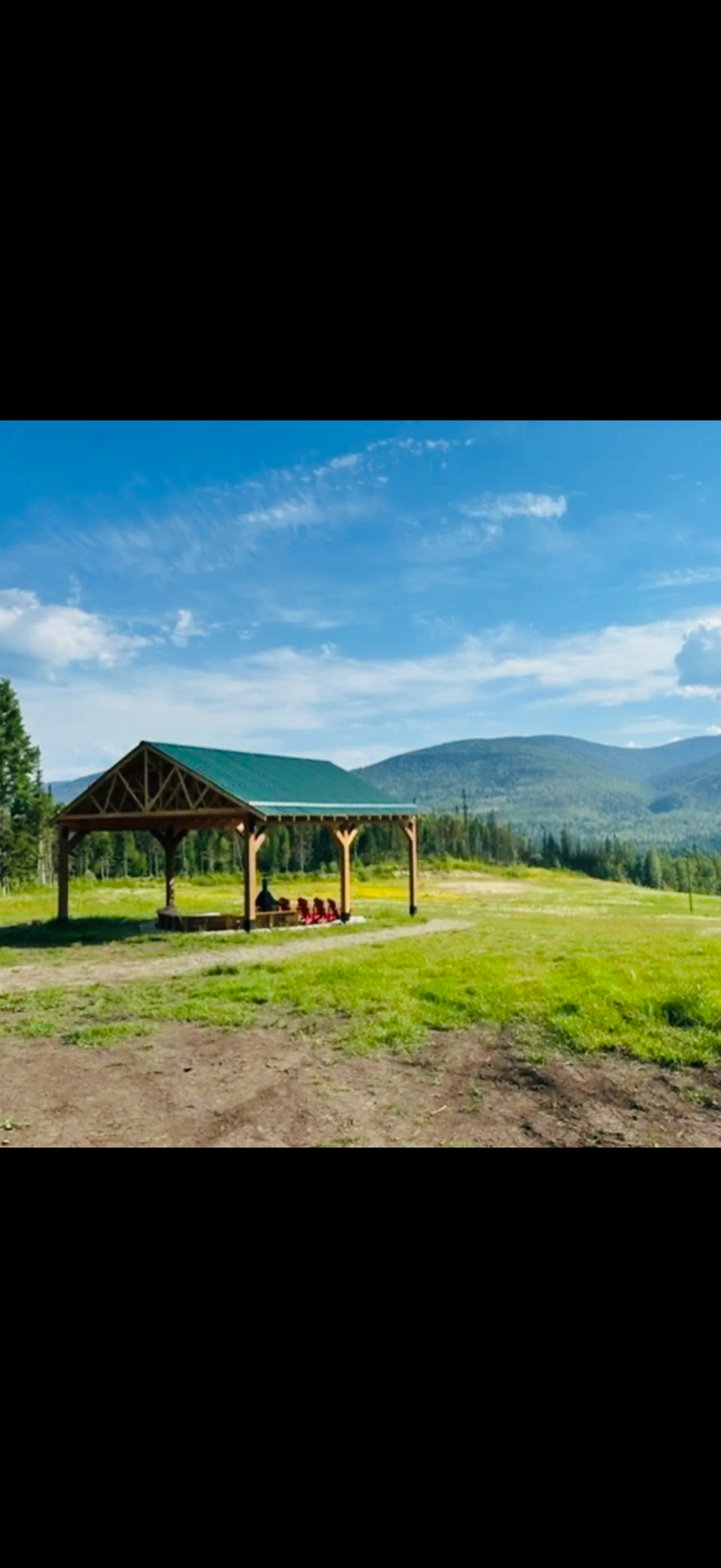 A rural outdoor scene featuring a wooden shelter with a green roof, set against a backdrop of rolling mountains and a blue sky with scattered white clouds.