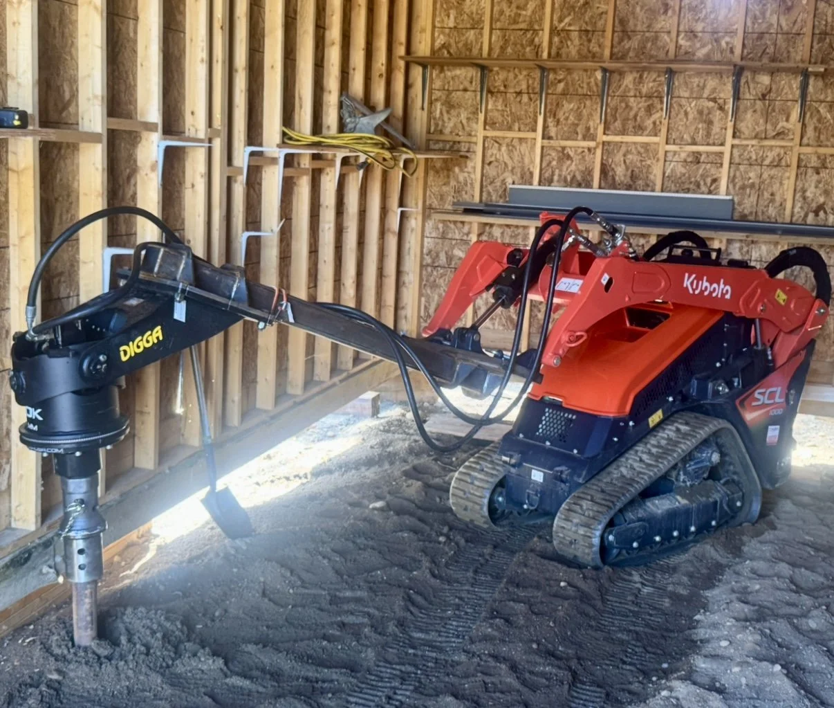 A small red Kubota tracked excavator with a Digga support arm attachment inside a wooden shed under construction.