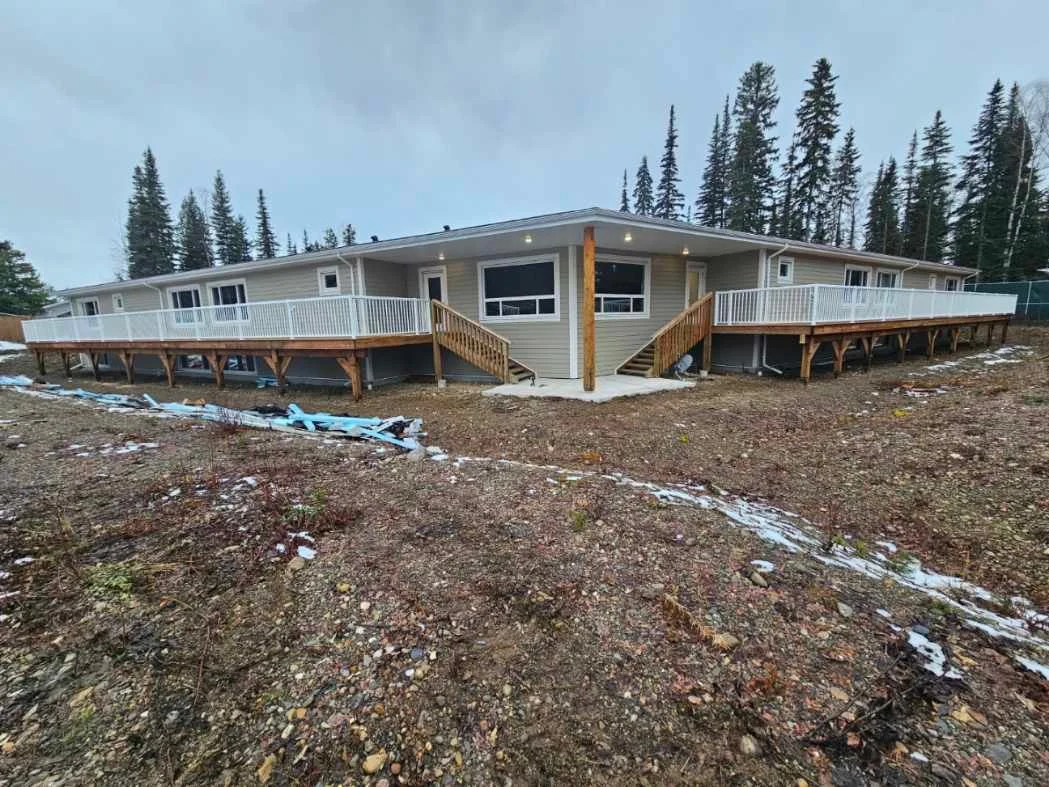 A single-story house on a hillside with a large wraparound deck, surrounded by pine trees, under a cloudy sky, with patches of snow on the ground.