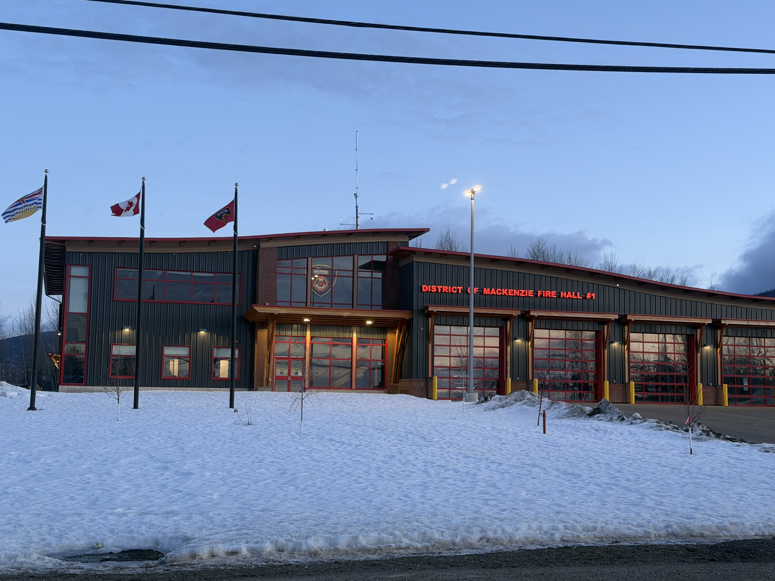 The Mackenzie Fire Hall #1 building during dusk, with snow on the ground, four flags on flagpoles, and illuminated signage reading 'District of Mackenzie Fire Hall #1' in red.