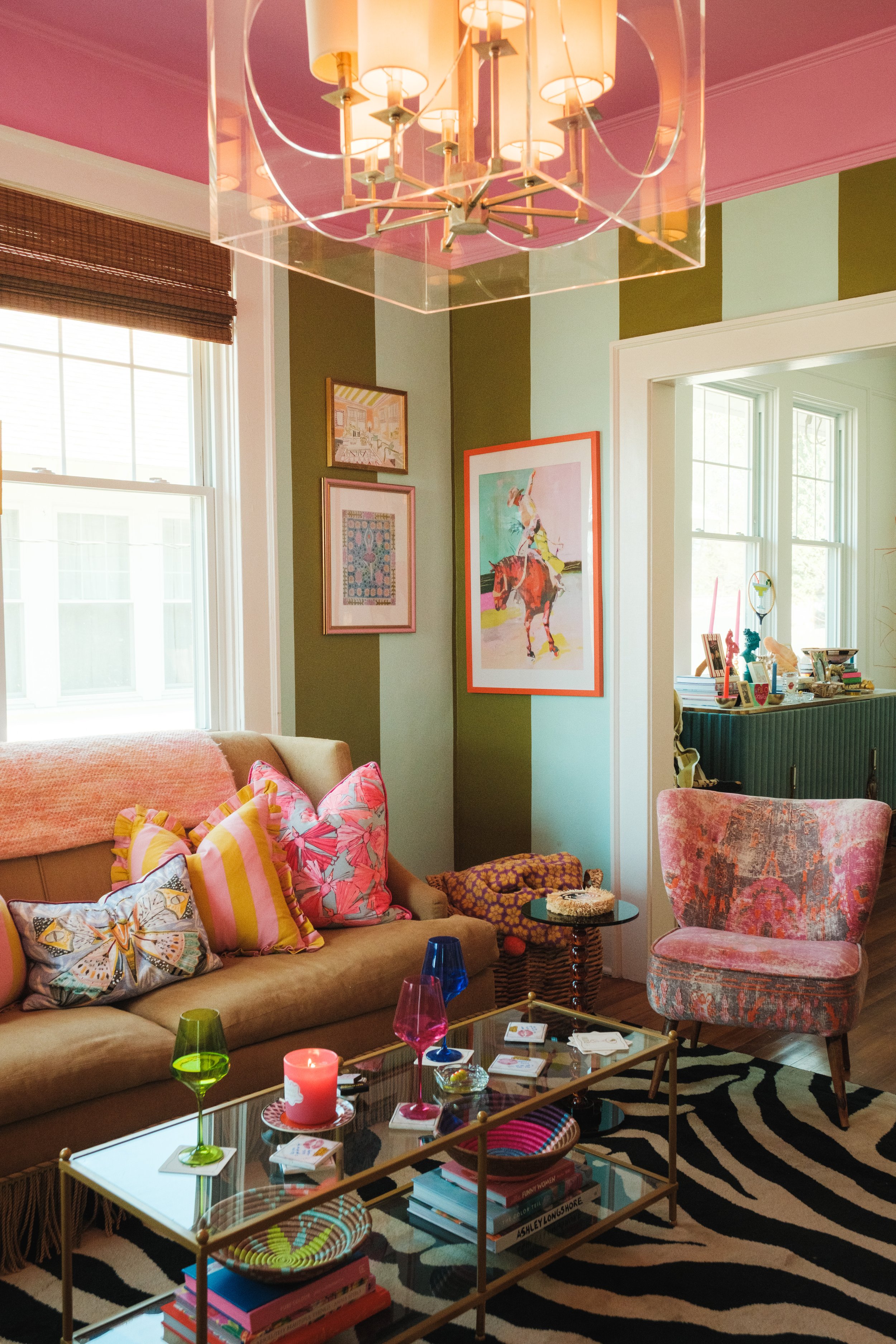 Living room with a beige sofa decorated with colorful pillows, a glass coffee table with candles, glasses, and books, a patterned armchair, and walls adorned with framed artwork. Large windows provide natural light, and a modern chandelier hangs from the ceiling.