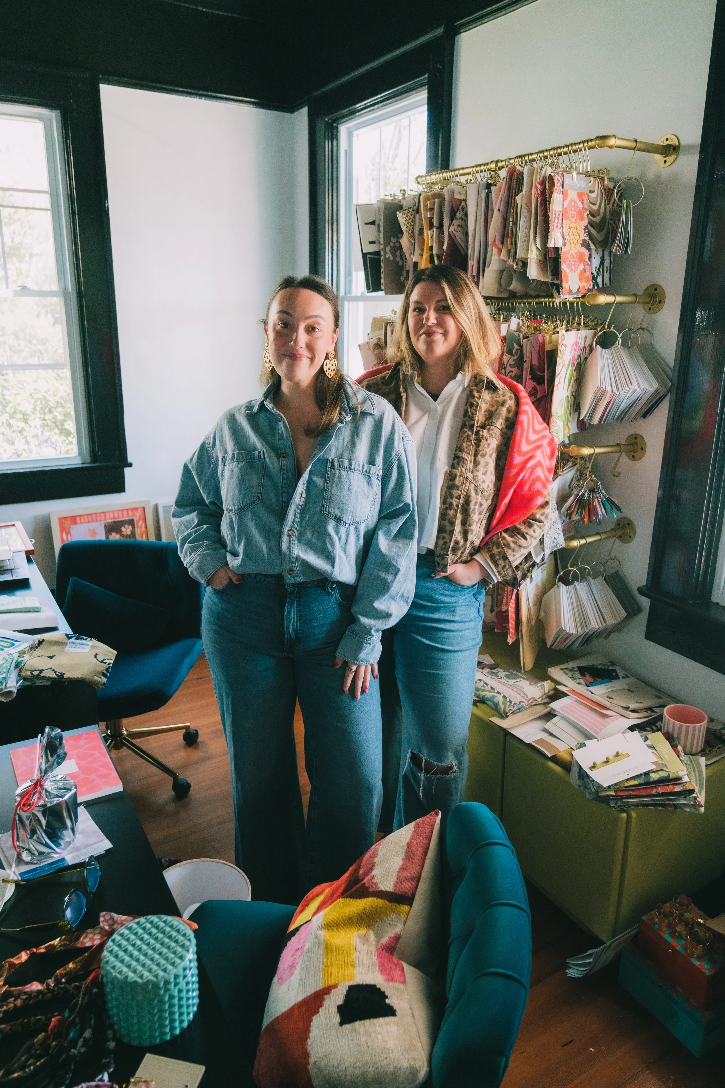Two women standing in a room filled with fabric swatches, craft supplies, and a desk, smiling at the camera.