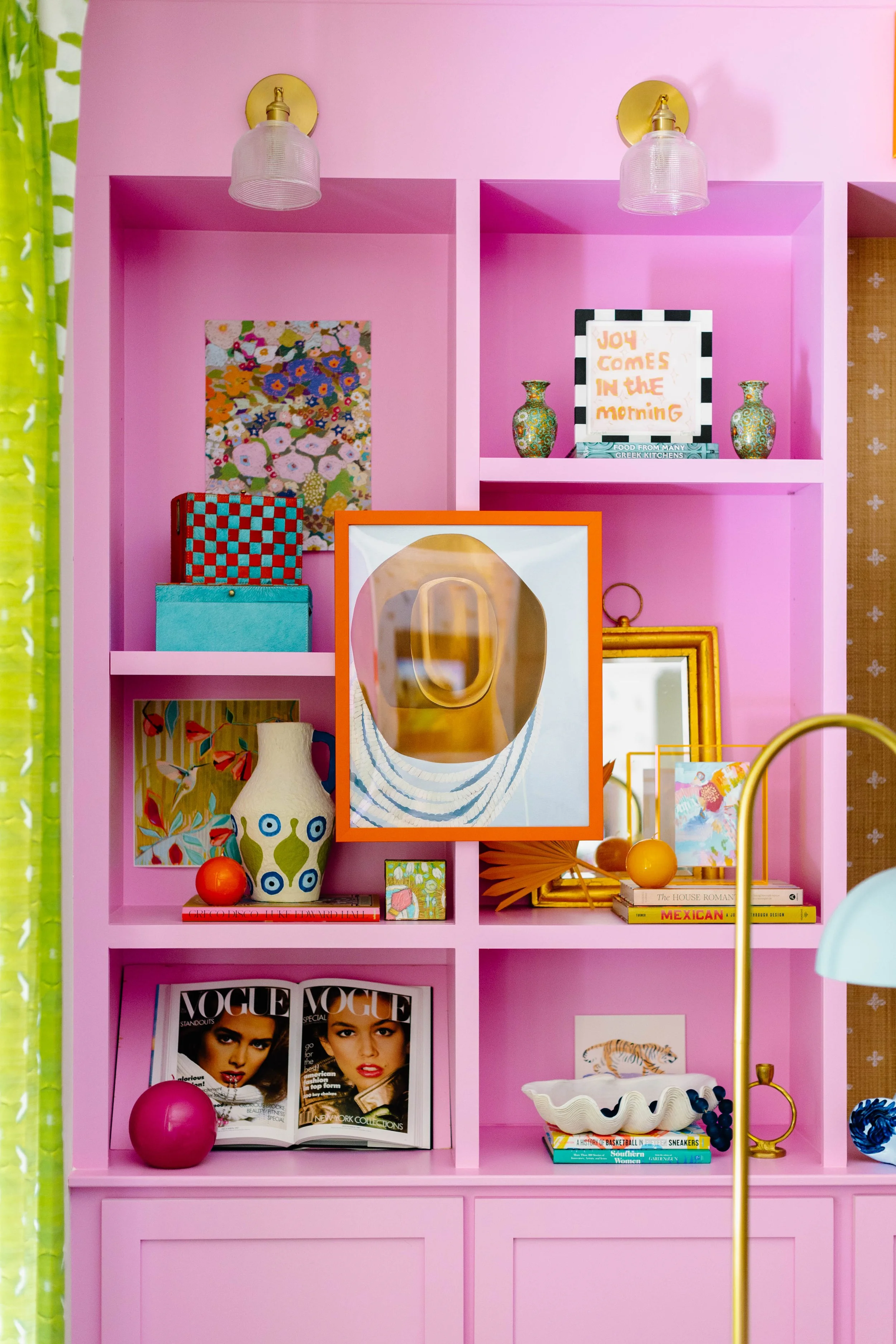 Pink bookshelf filled with colorful decor, books, and magazines, including Vogue, with decorative vases, artwork, a framed quote, and various small objects.