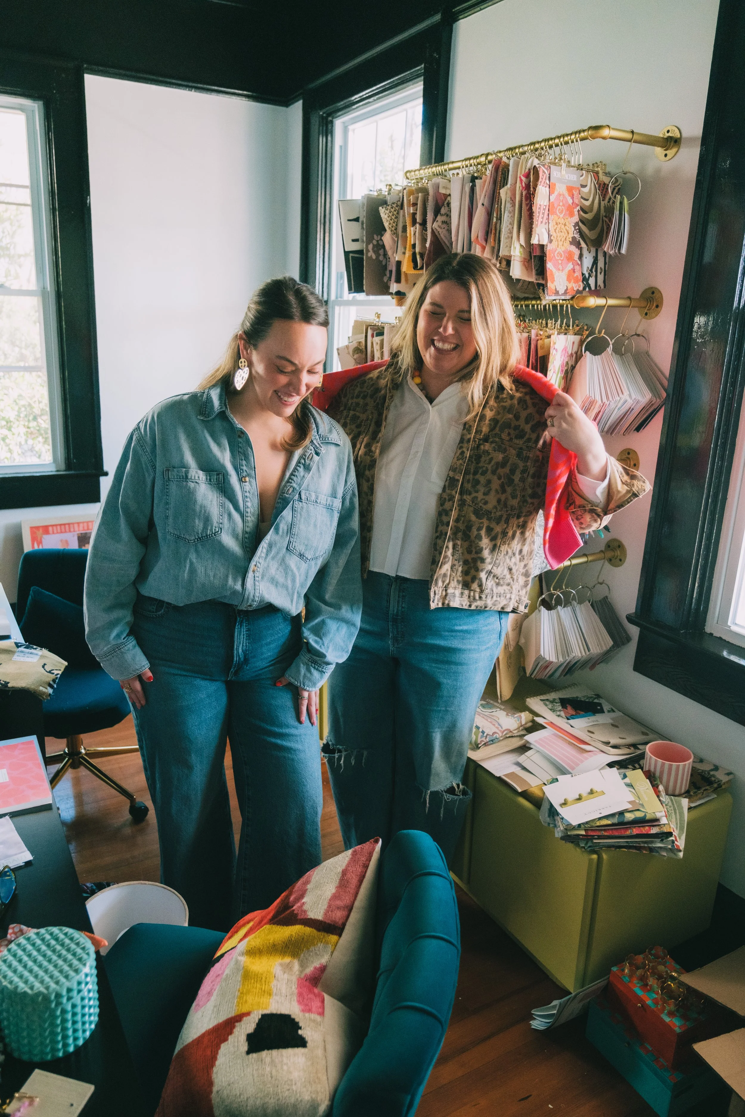 Two women are smiling and looking down in a colorful, well-decorated room with fabric samples and design materials, suggesting a creative or design workspace.