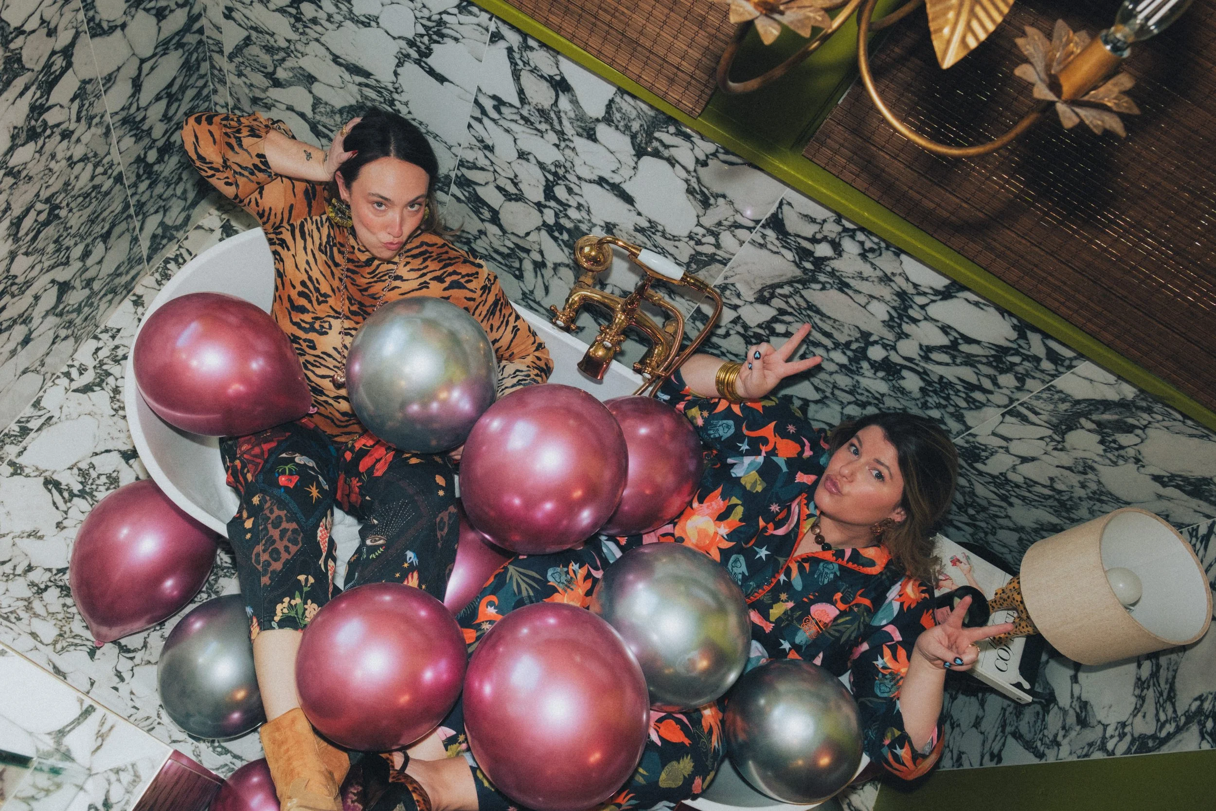 Two women in colorful pajamas posing in a bathtub filled with pink and silver balloons, in a room with black, white, and green marble walls and a gold faucet.