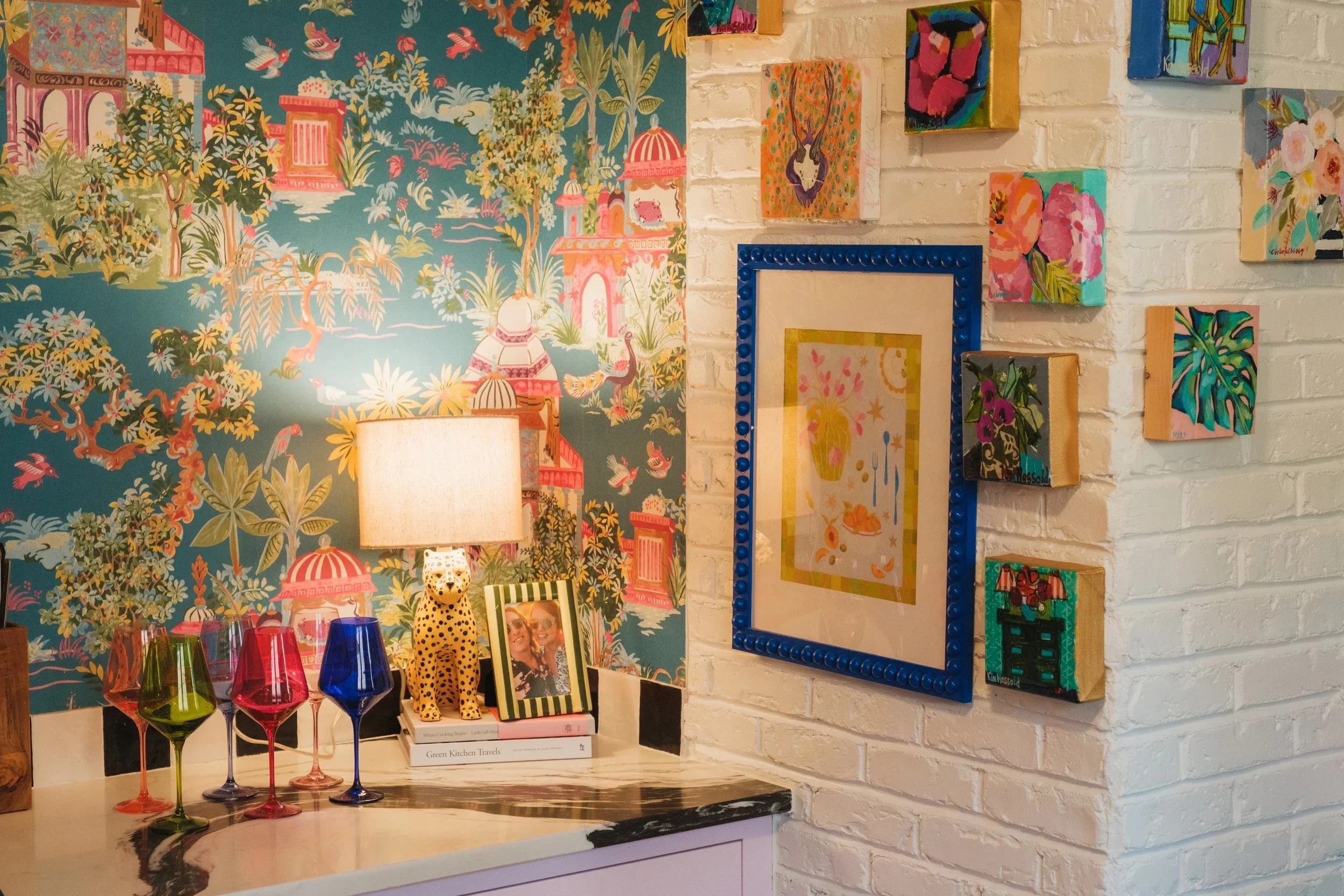 Decorative interior wall with colorful artwork, a table with glasses, a lamp, a framed photo, and a playful leopard lamp. One wall has blue floral mural, and the other has hanging framed pictures on a white brick wall.