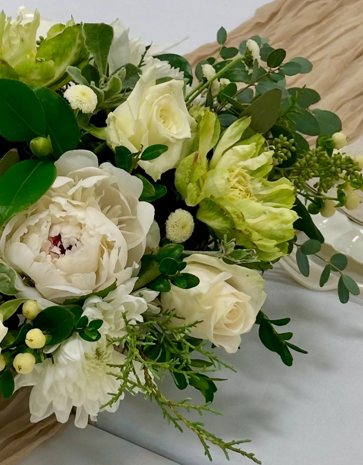 Close-up of a bouquet of white and cream flowers including roses, peonies, and lilies, with green foliage, on a table.